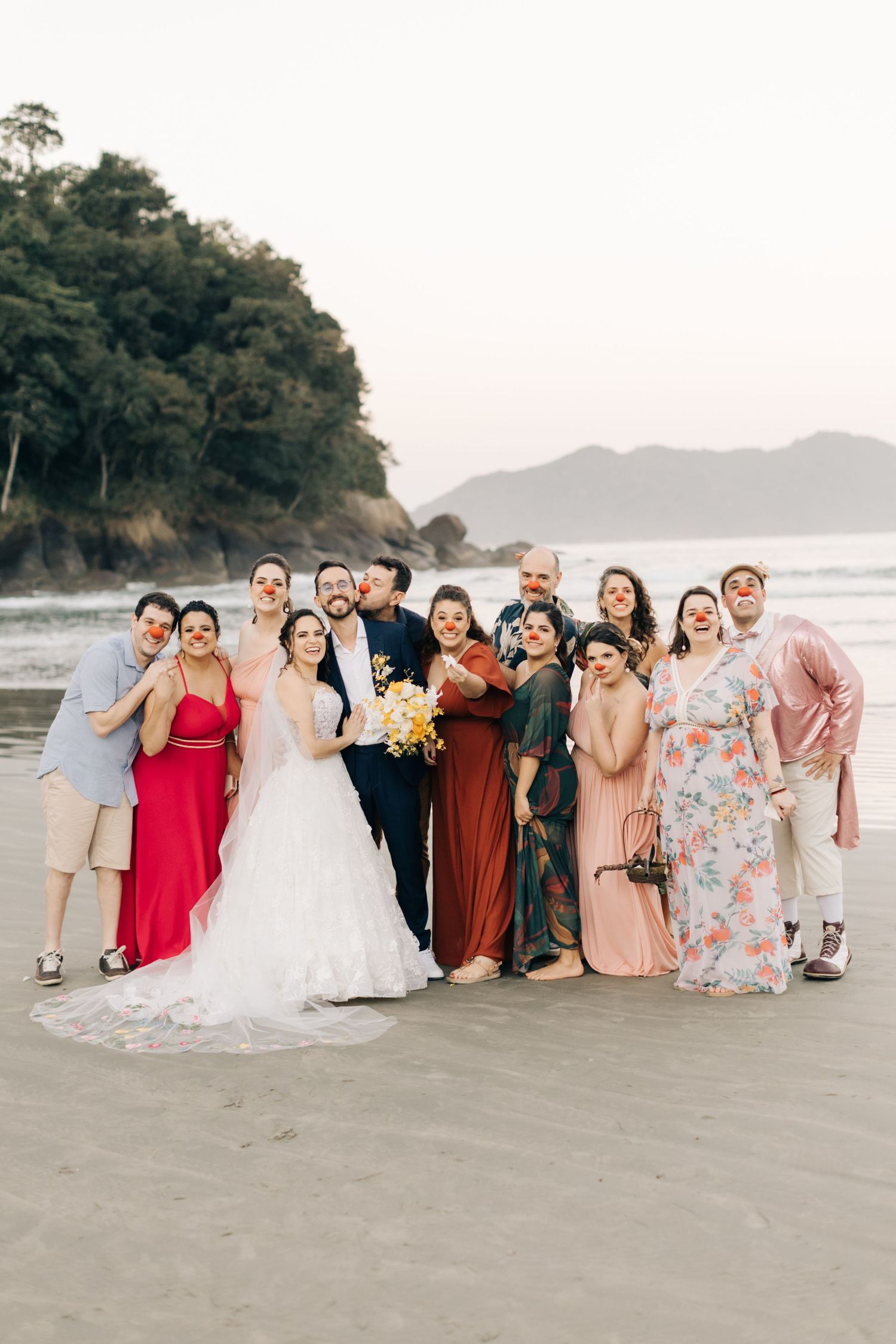 Wedding party in colorful attire poses together on a beach with mountains and forest in the background.