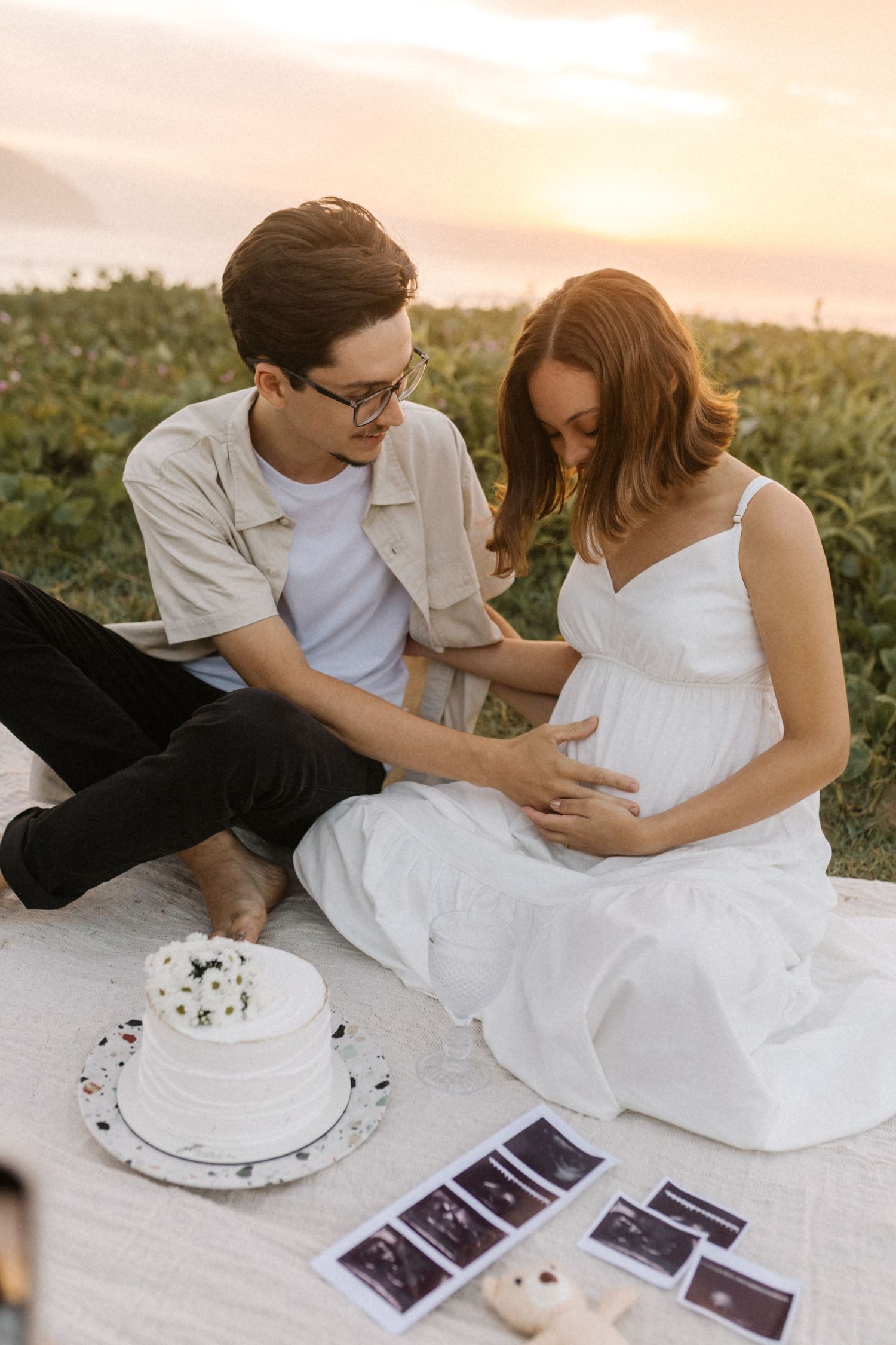 A couple shares an intimate moment while looking at ultrasound photos and a small cake during a pregnancy announcement session.