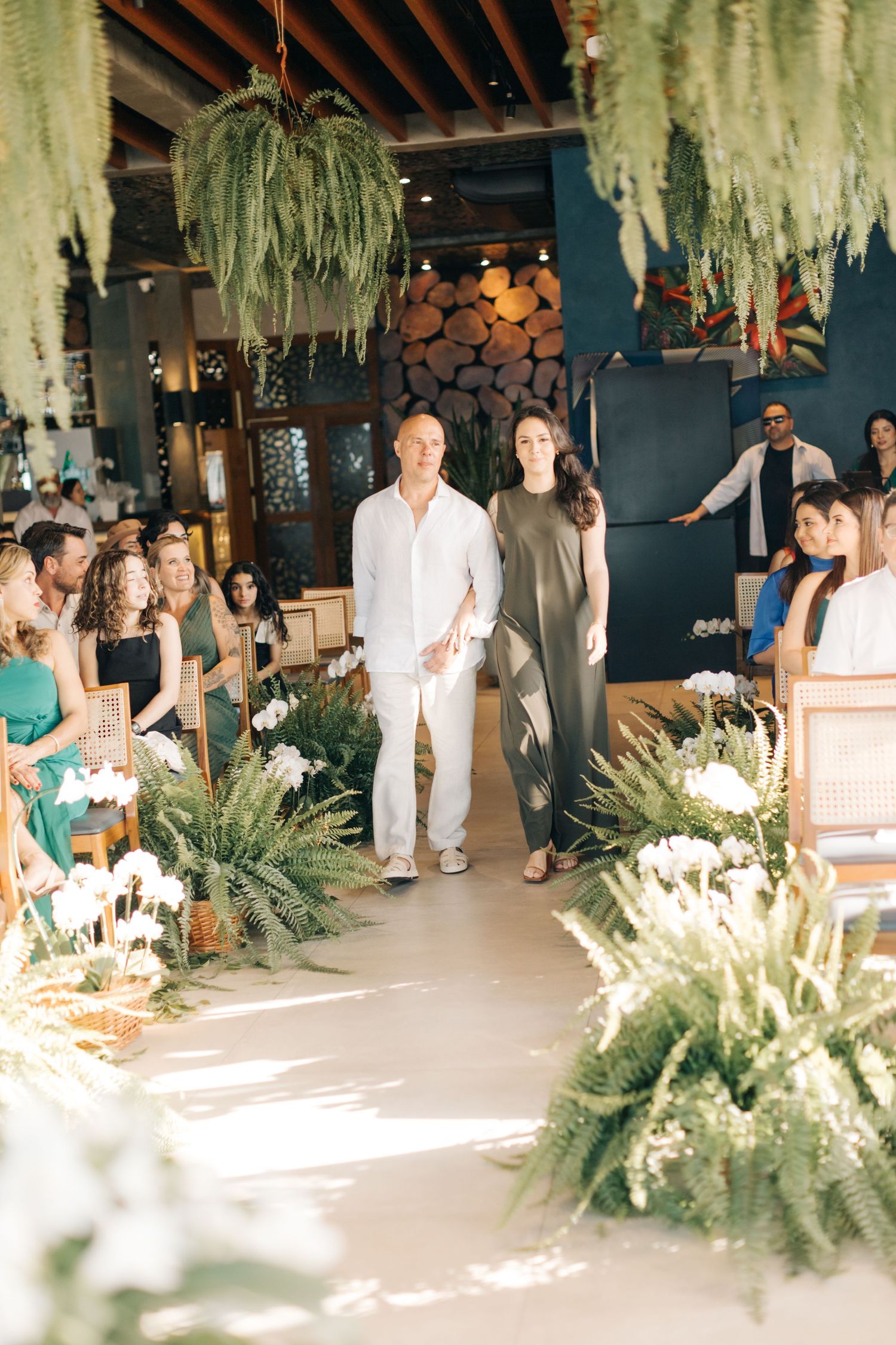Couple walking down aisle decorated with cascading plants and white flowers.