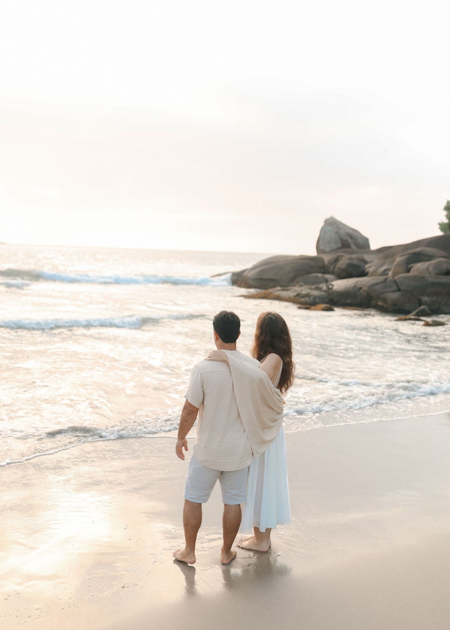 A couple stands together watching the sunset on a beach with rocky formations in the background.