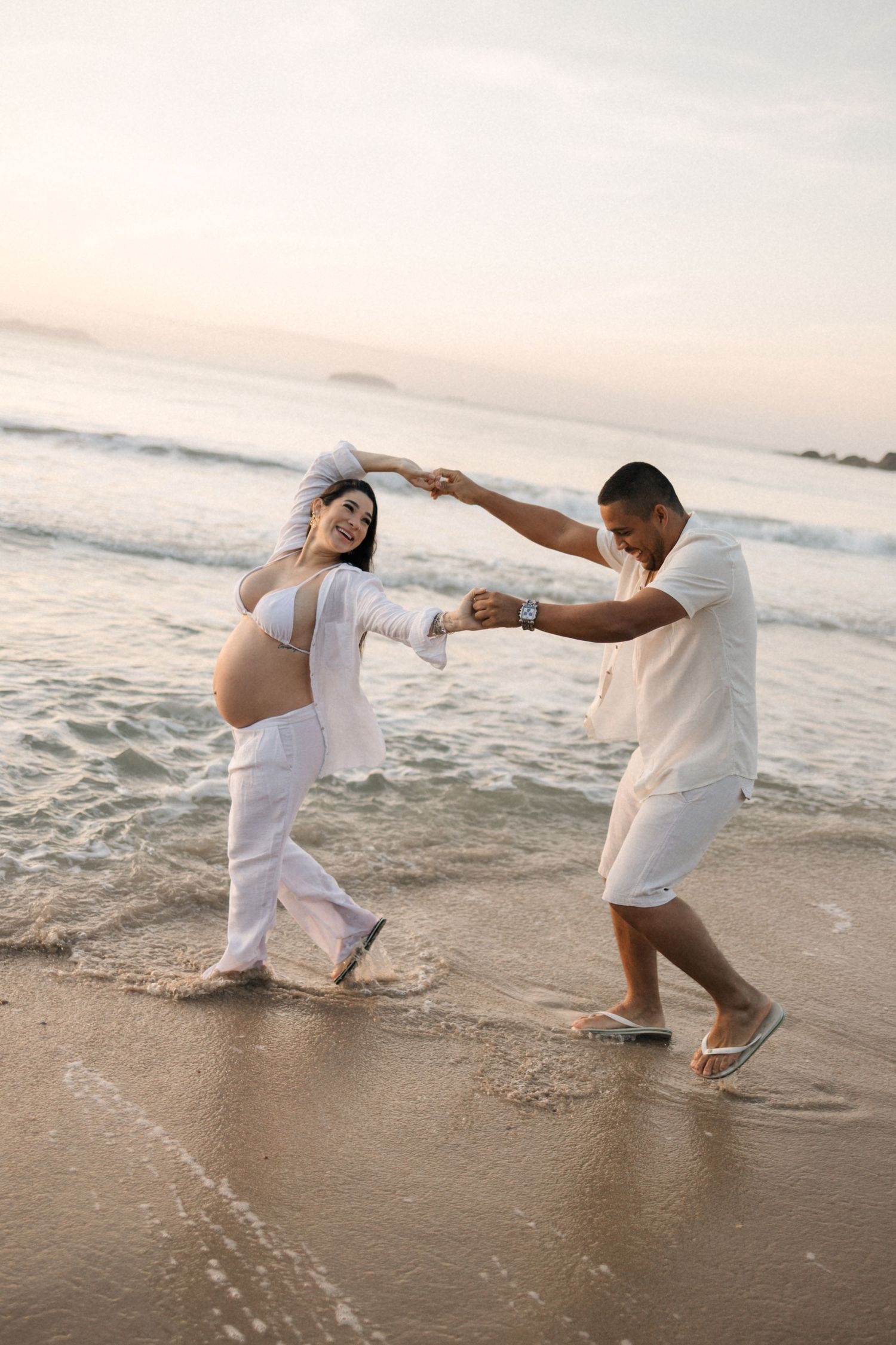 Dancing couple in white clothing enjoying playful moment on beach at sunset.