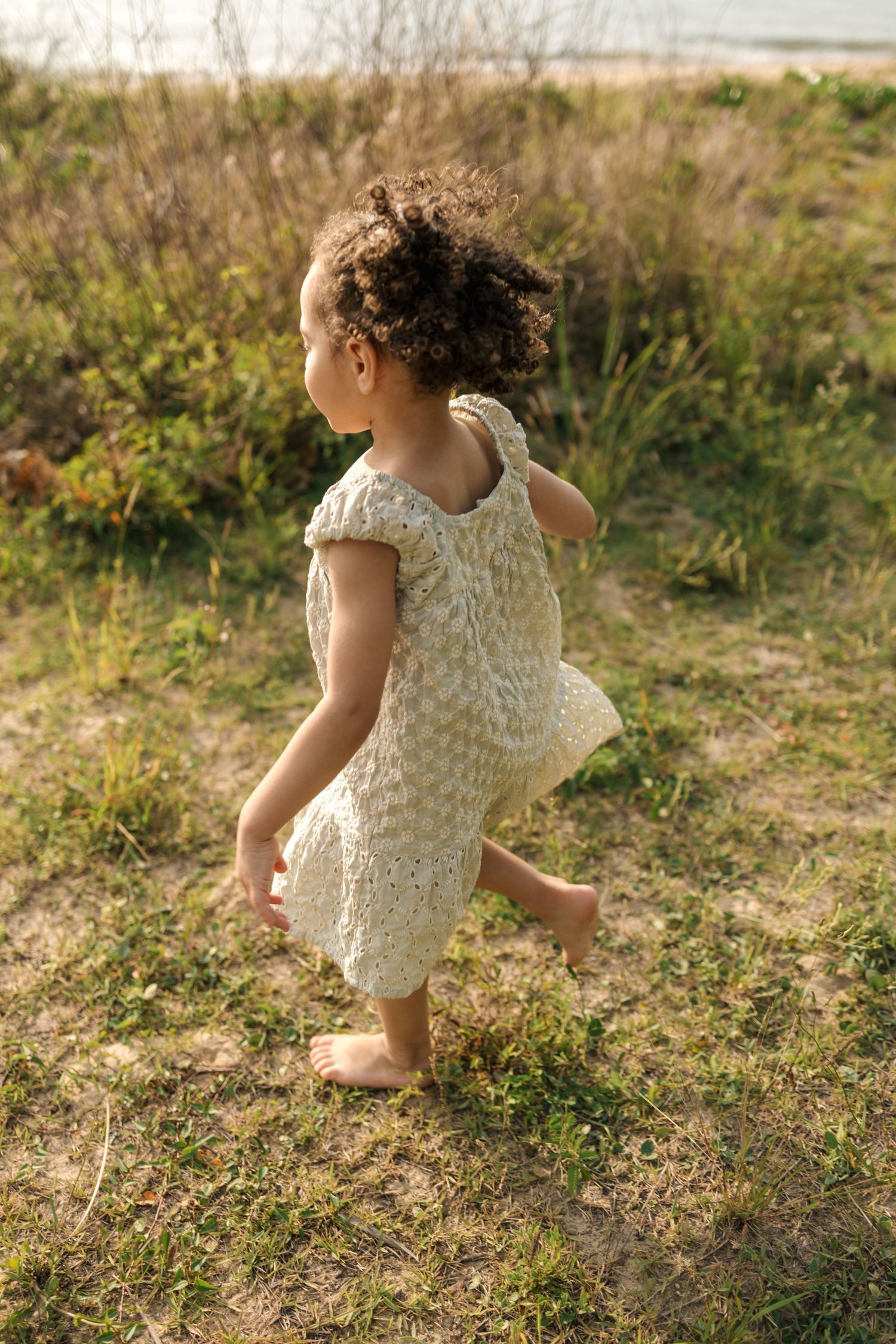 A young child in a white lace dress dances in a sunlit meadow with wild grasses and flowers.