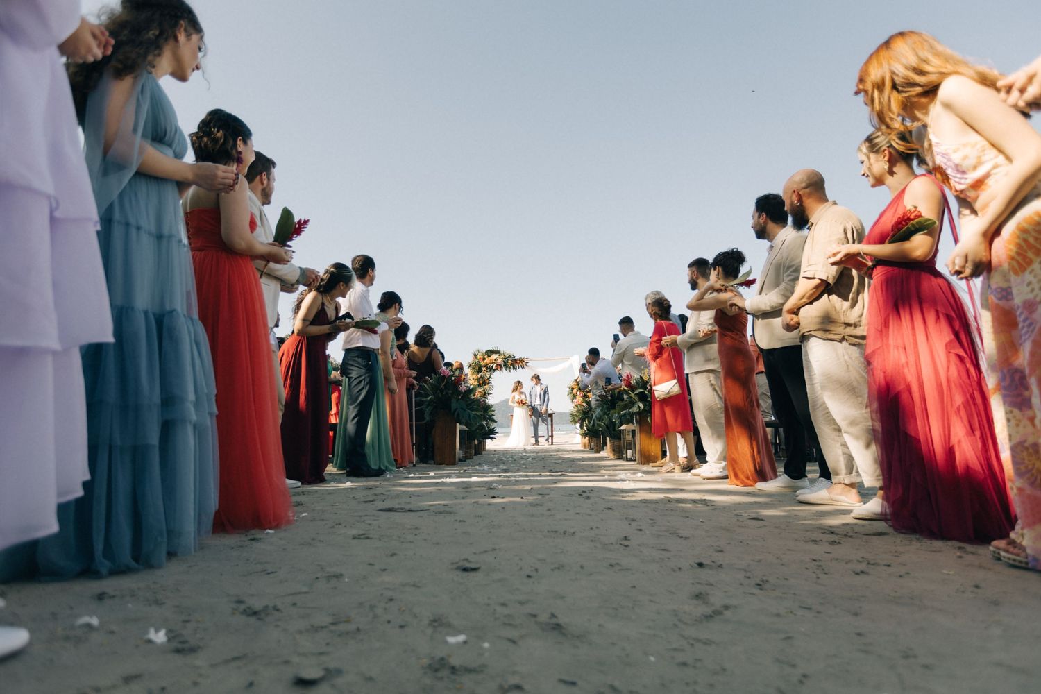 Wedding guests form pathway on beach holding phones to capture ceremony moments.