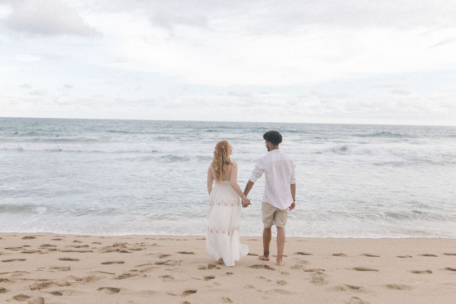A couple walks together on a sandy beach with ocean waves in the background.
