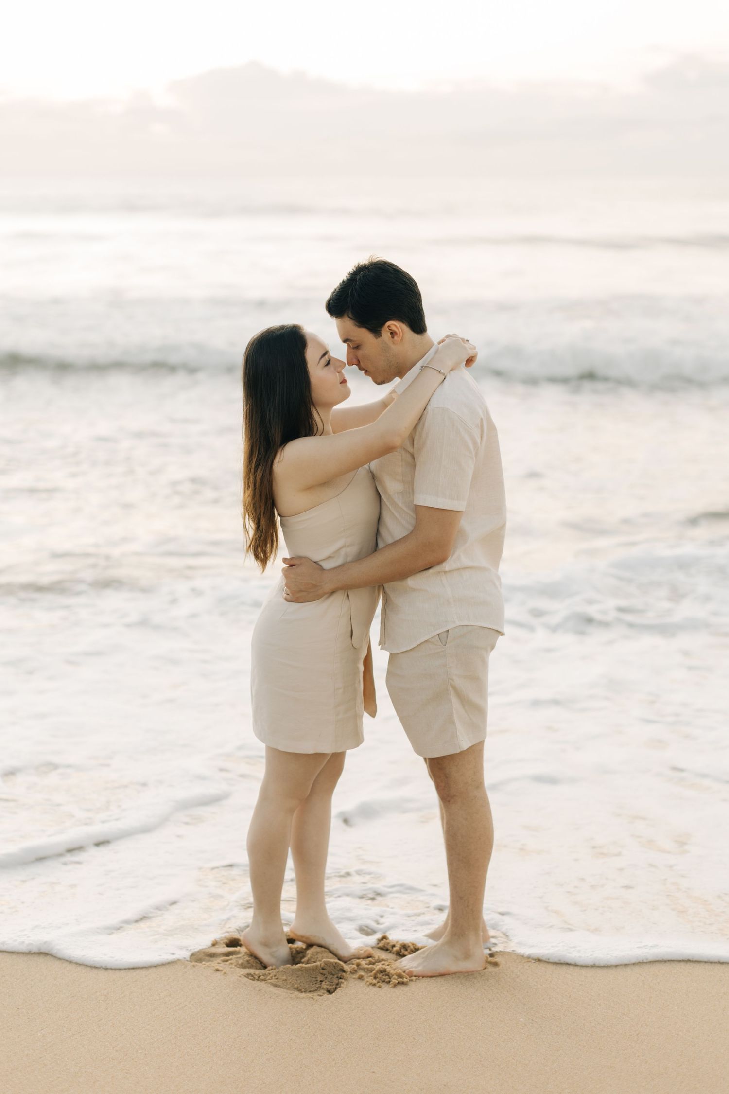 Romantic couple embracing on a beach at sunset wearing matching light colored outfits.