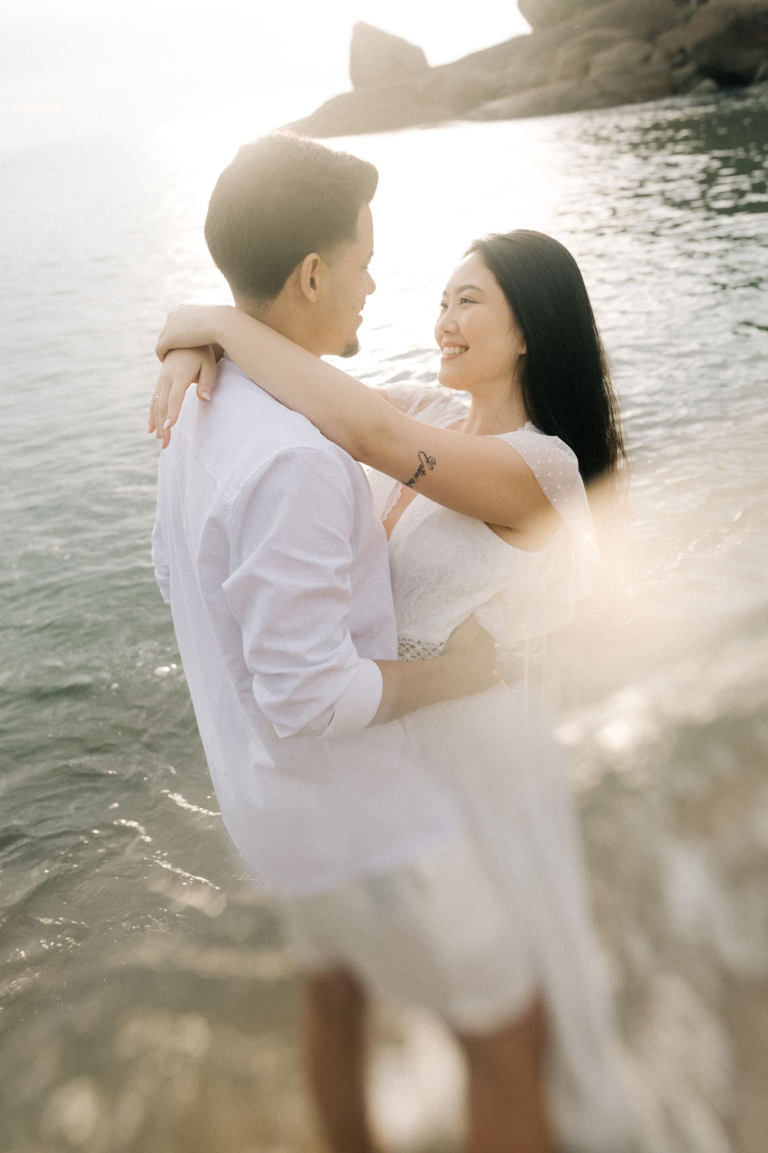Romantic couple in white outfits embrace on a sunlit beach during golden hour.