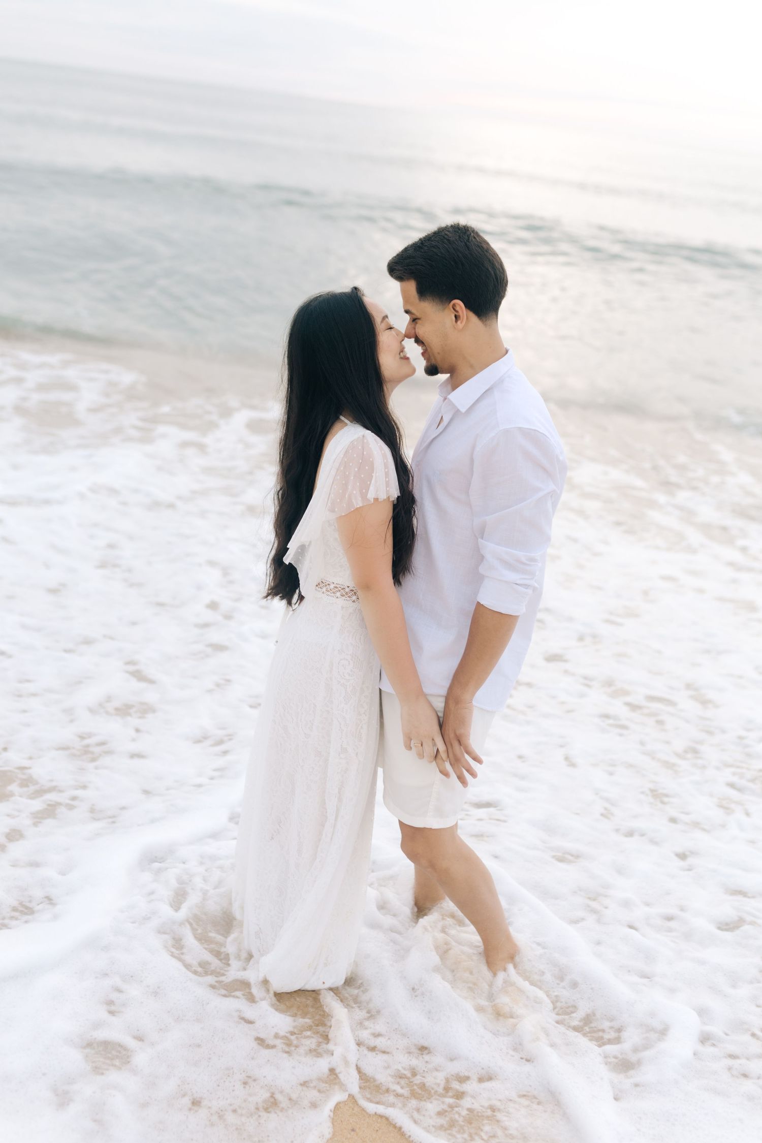 A tender moment shared between a couple in white attire on a beach at sunset.