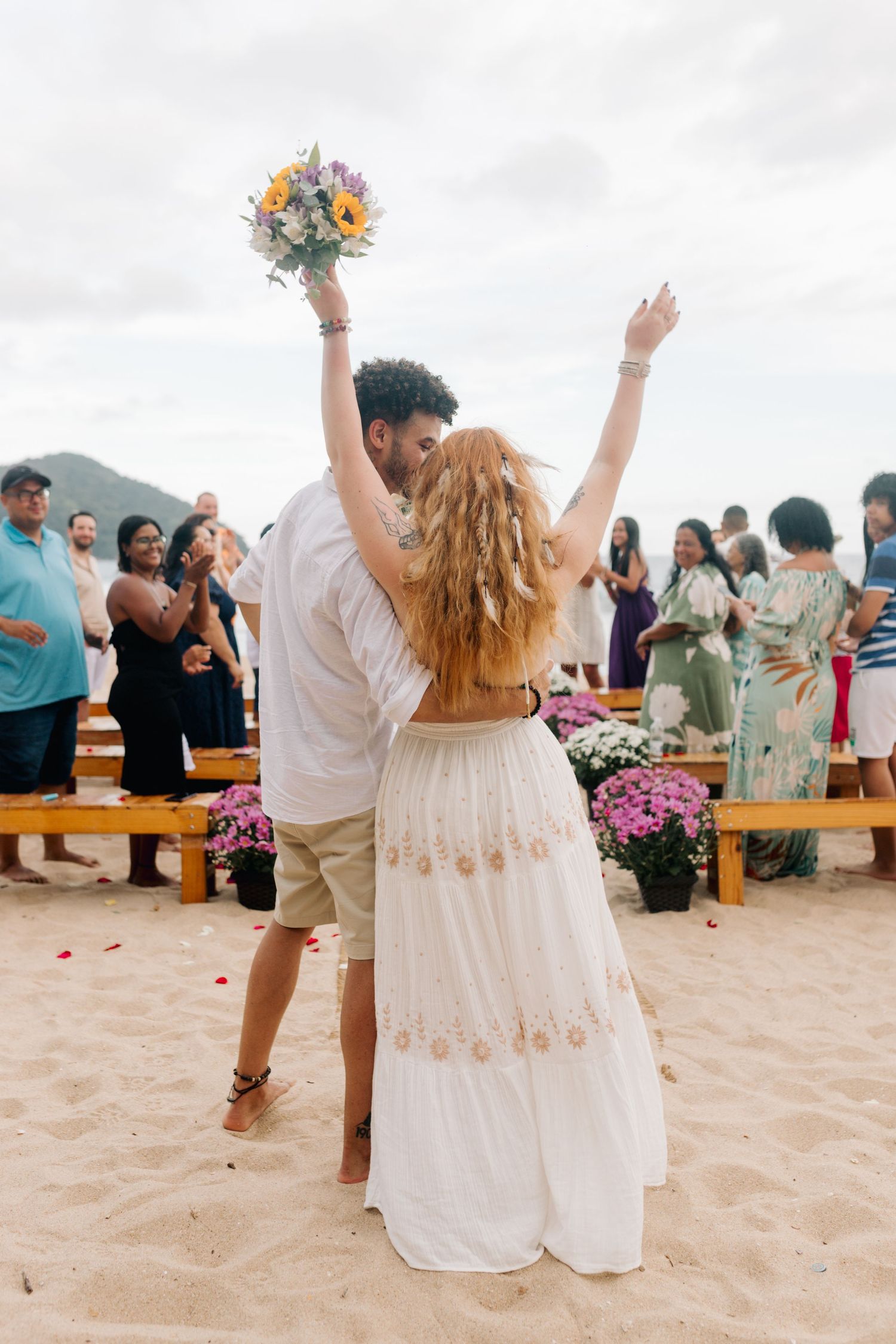 A celebratory moment as newlyweds raise their arms in joy on a beach surrounded by pink flowers.