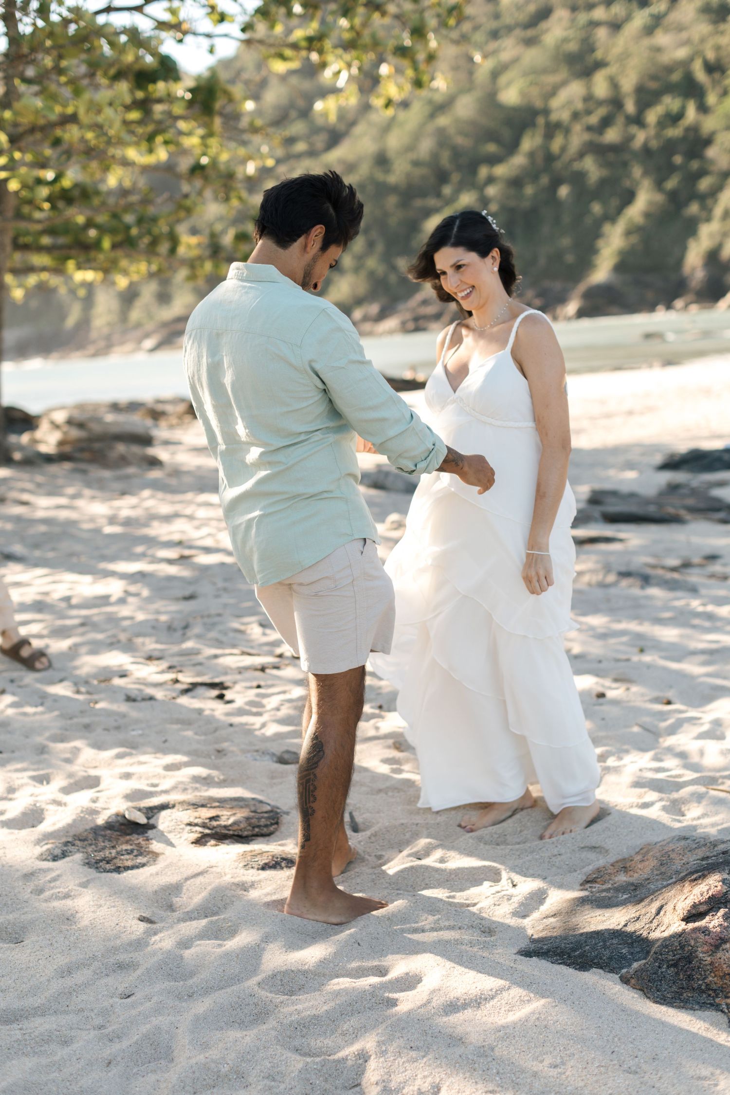 A couple in white and mint attire dances playfully on a sandy beach during a photo session.