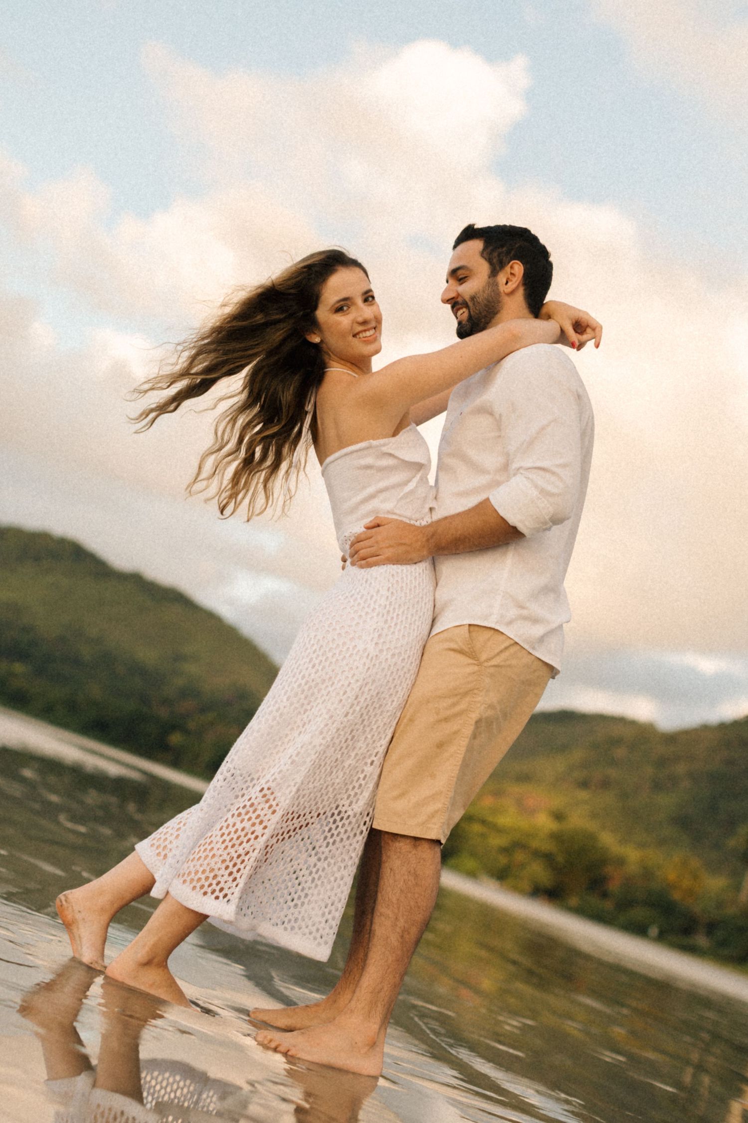 Dancing couple in white dress and khaki shorts enjoying sunset moment on beach.