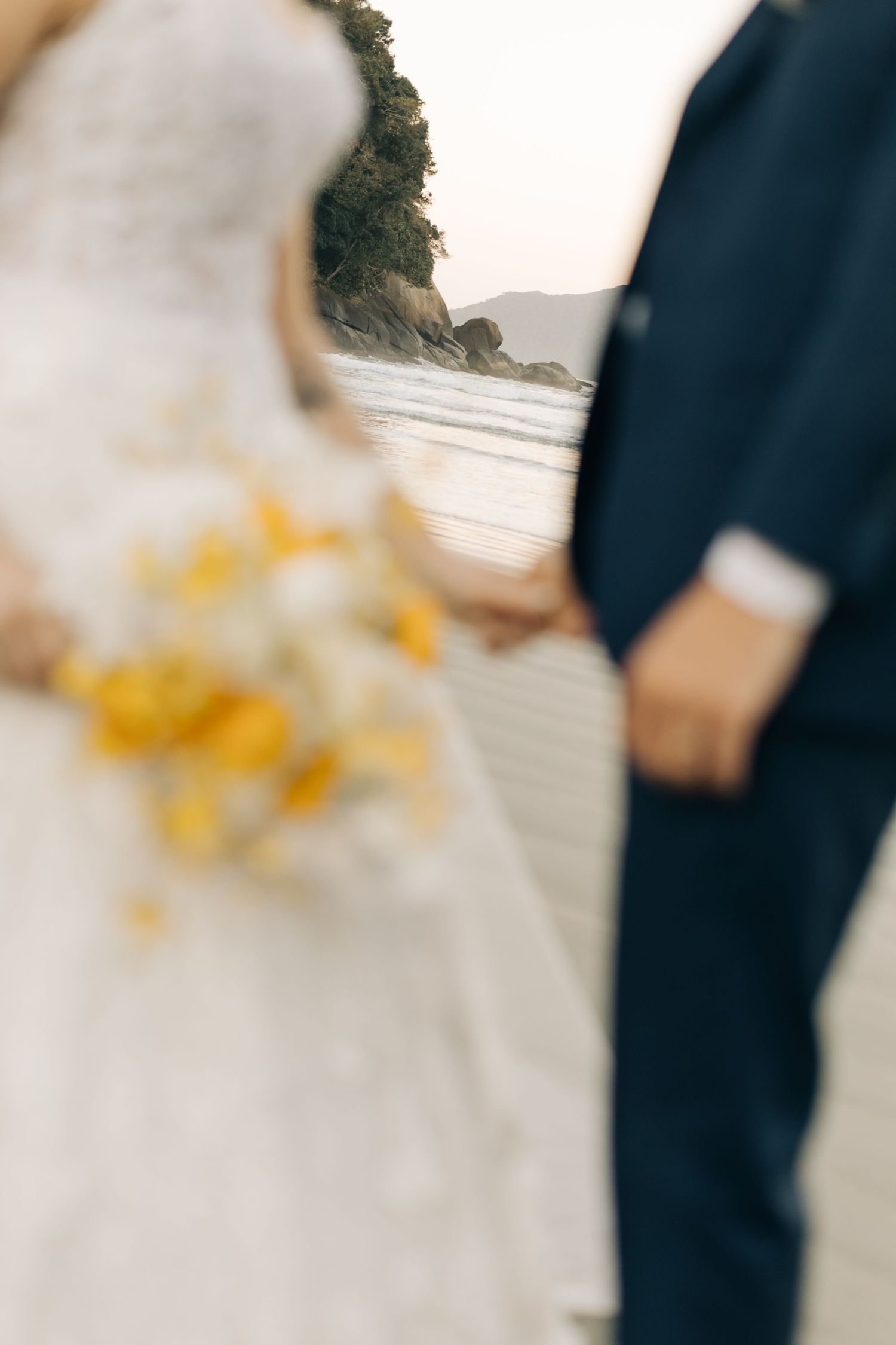 Close-up details of a wedding dress and bouquet featuring yellow flowers on the beach.