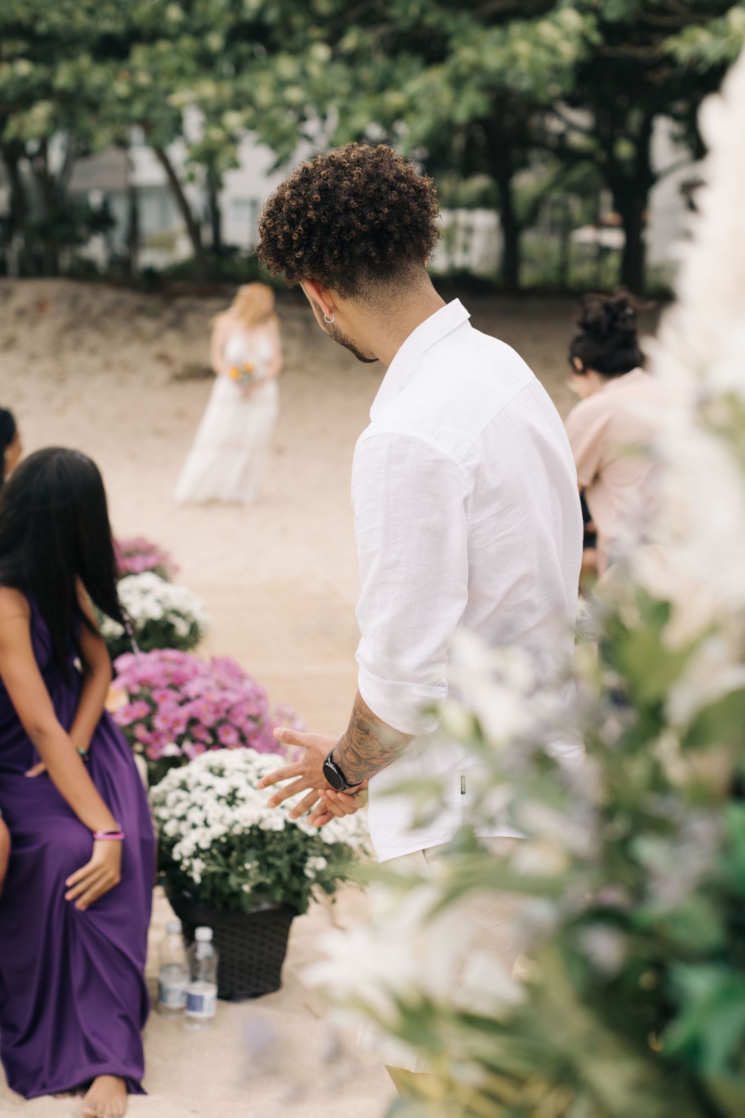 Wedding guest in white shirt standing among purple-dressed attendees and floral arrangements.