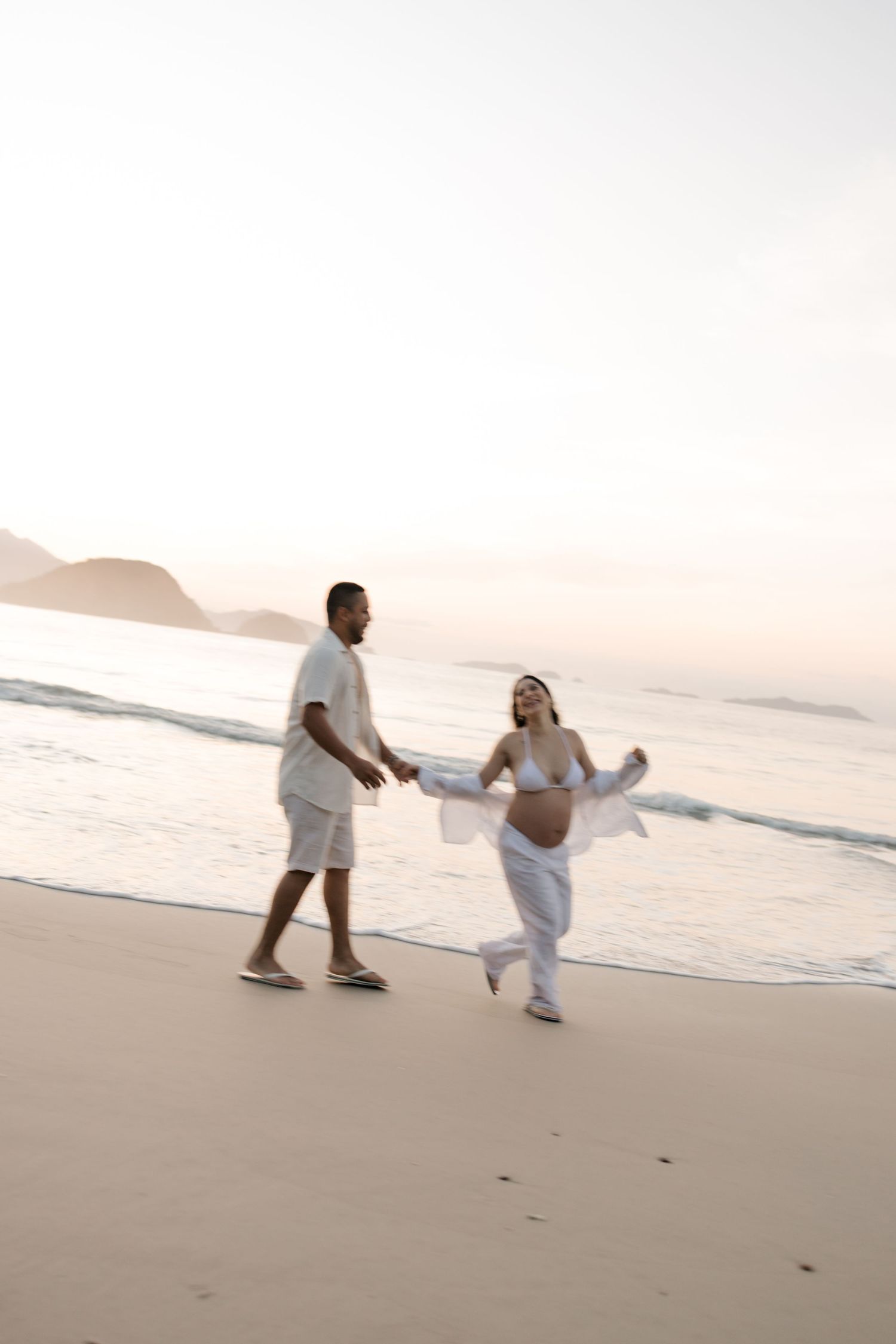 Couple in white clothing dancing together on beach during maternity photo session.