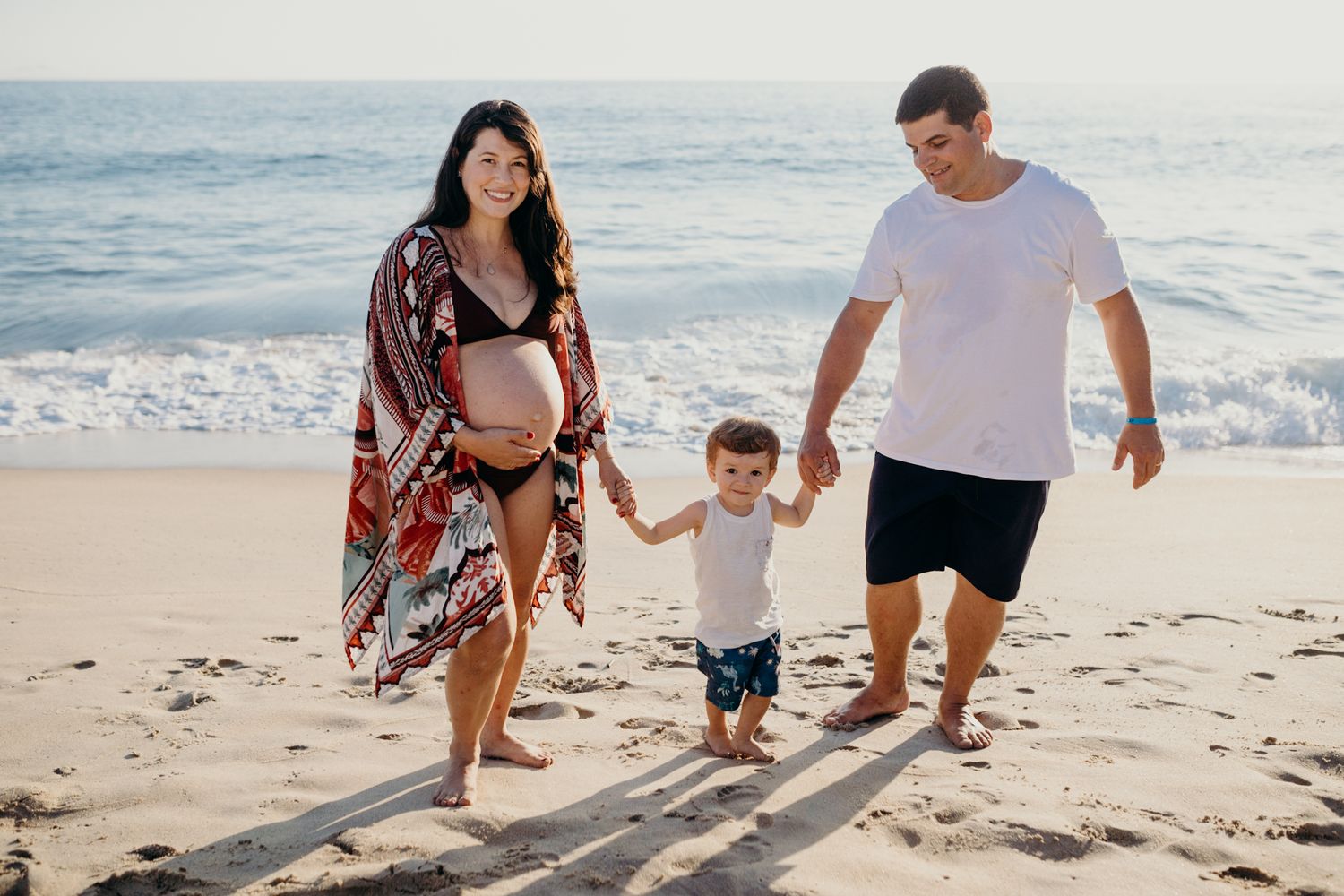 Family walking hand-in-hand along a sunny beach with ocean waves in the background.