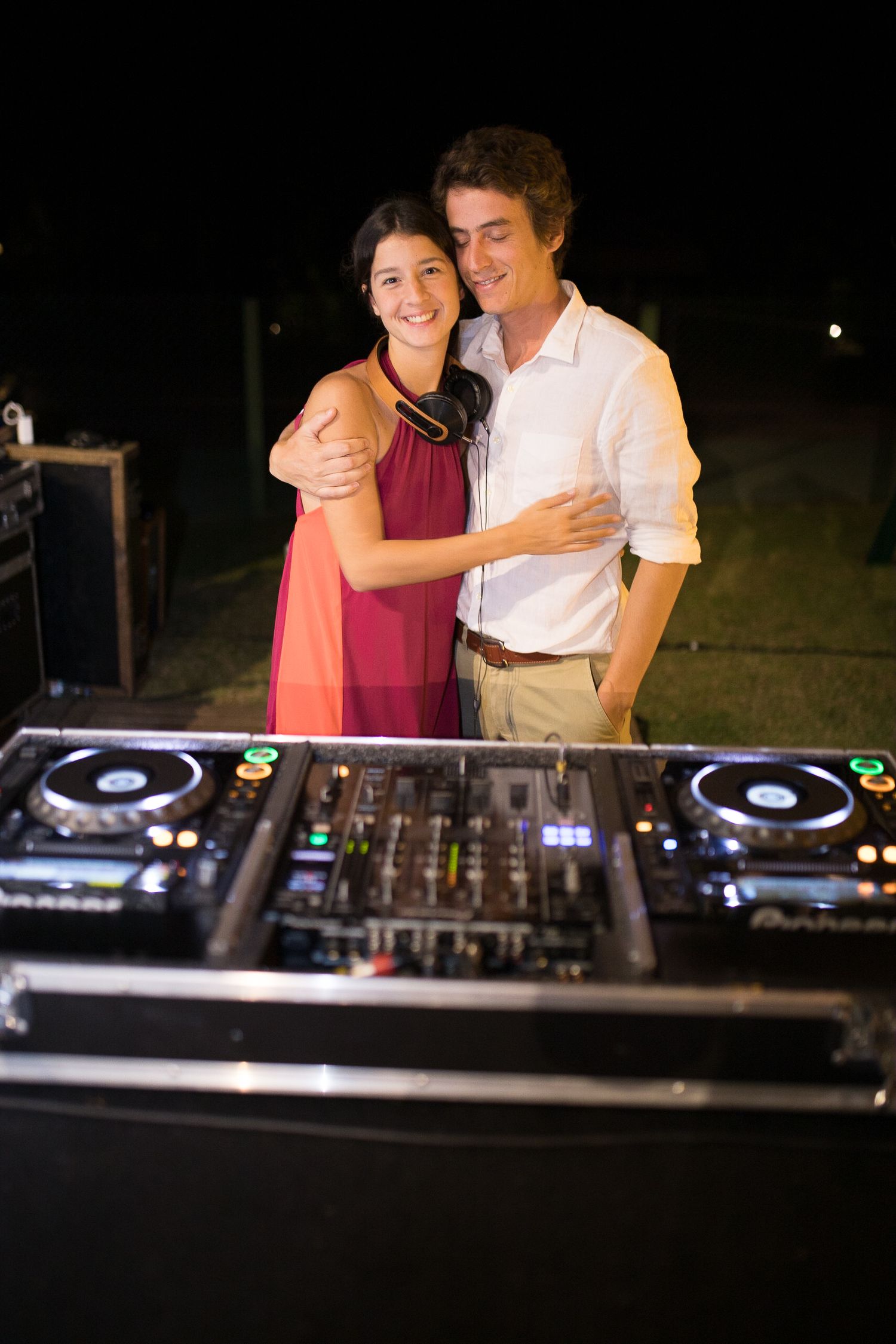 Couple poses behind professional DJ equipment at outdoor wedding reception.