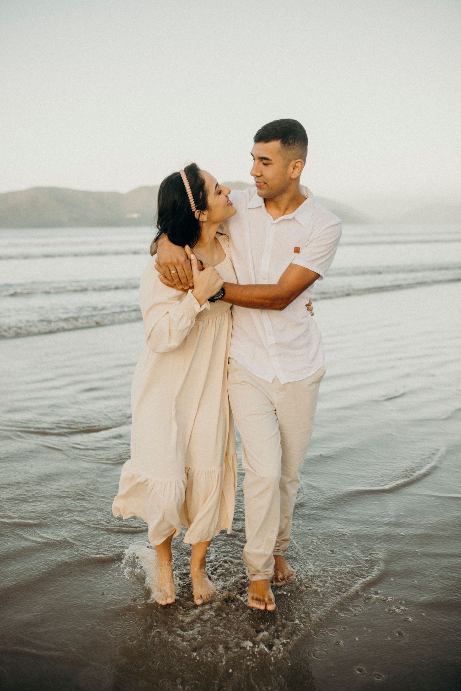 A couple in cream and white outfits dances and splashes in the shallow ocean waves.