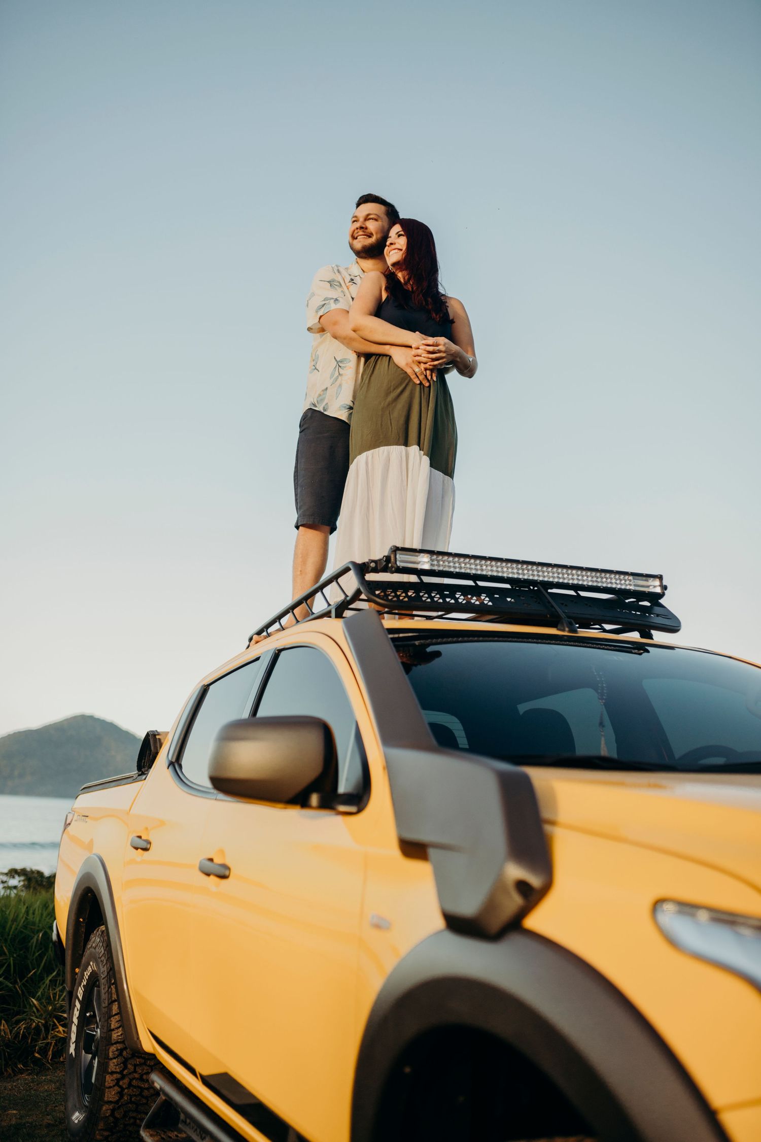 Couple embracing on roof of yellow pickup truck with mountain coastline in background.