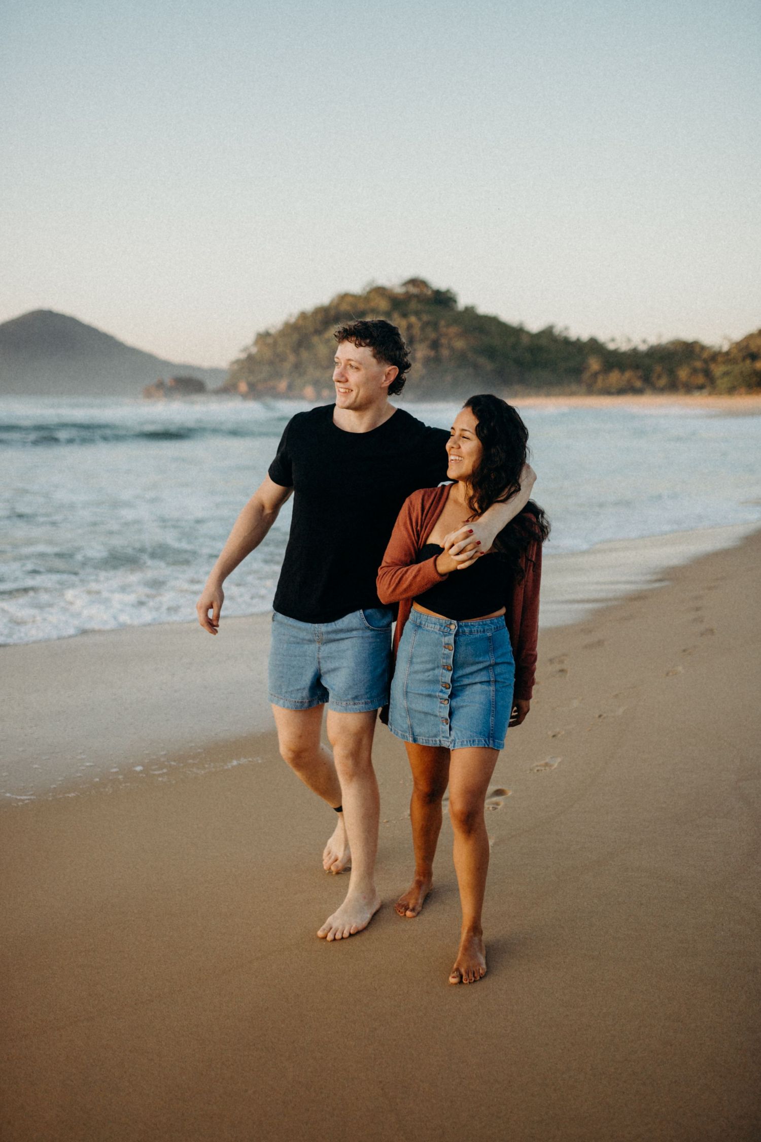 Couple strolling hand in hand on a sandy beach with mountains and ocean waves in background.