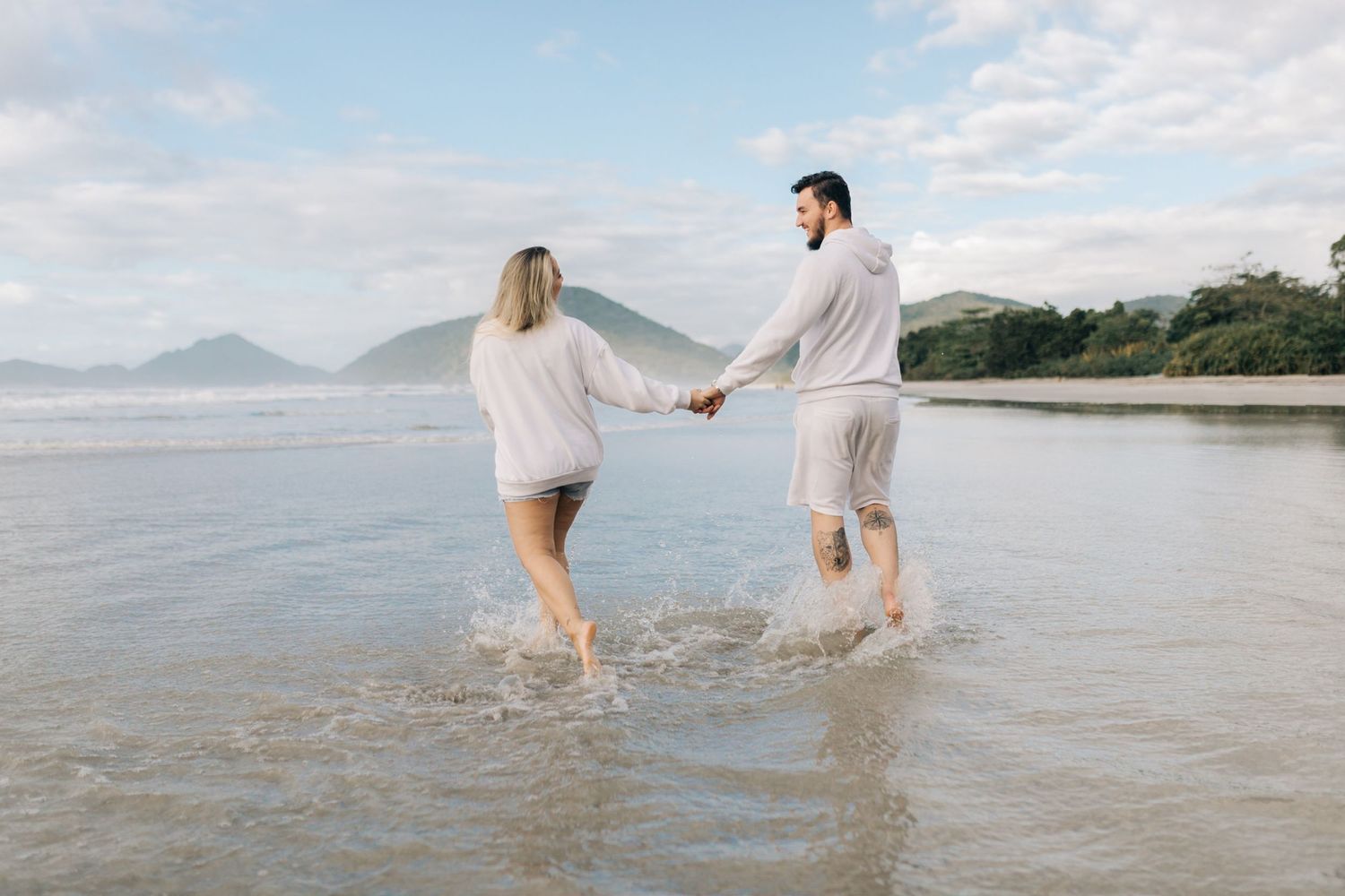 Couple splashing in ocean water with mountains and blue sky in background.