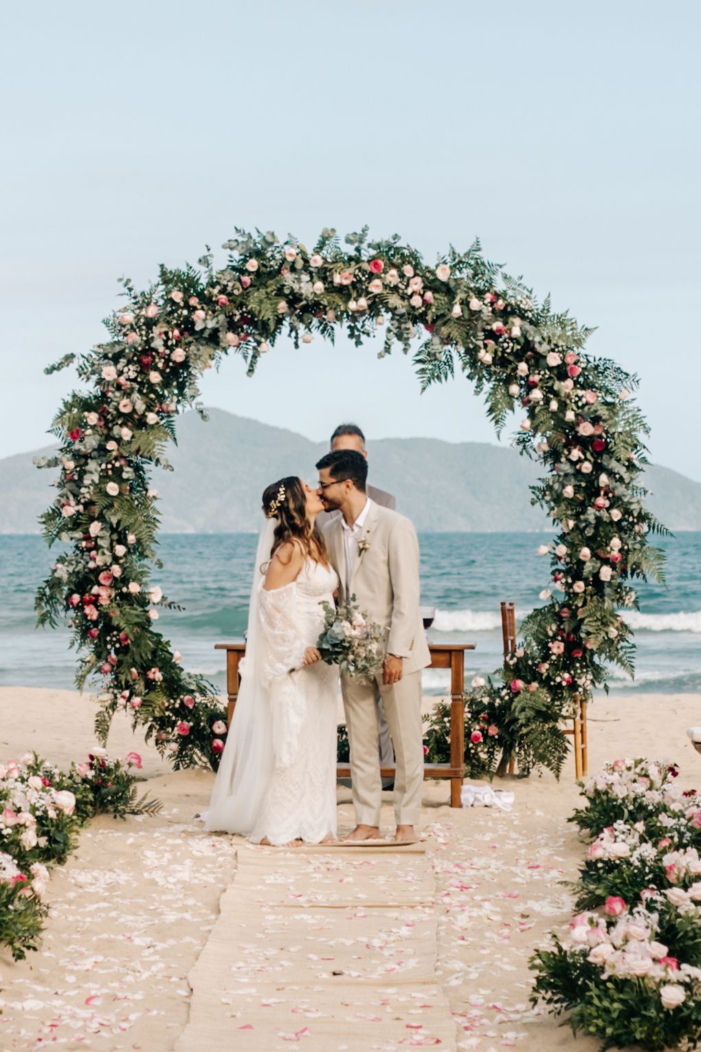 A wedding ceremony under a floral arch on the beach with mountains in the background.