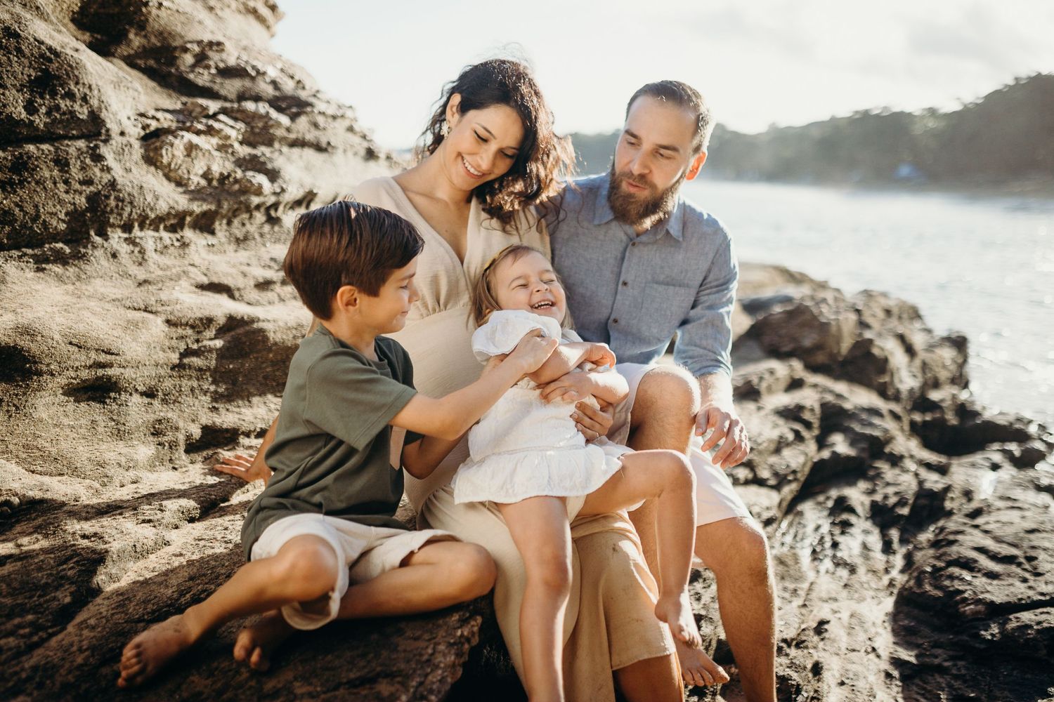 A tender family moment captured against a backdrop of coastal rocks and blue ocean waters.