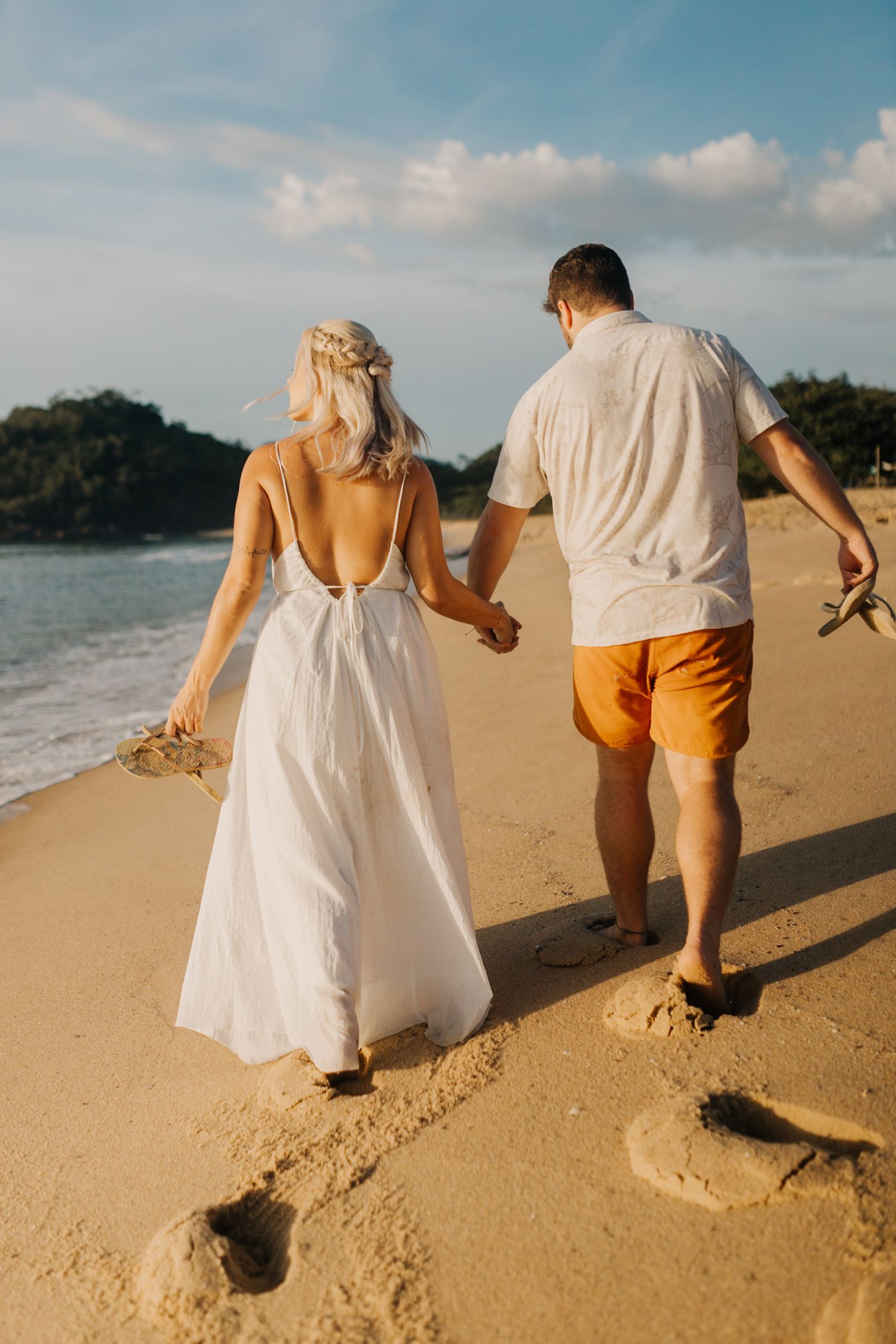 Couple walking hand in hand on sandy beach at sunset wearing white dress and orange shorts.