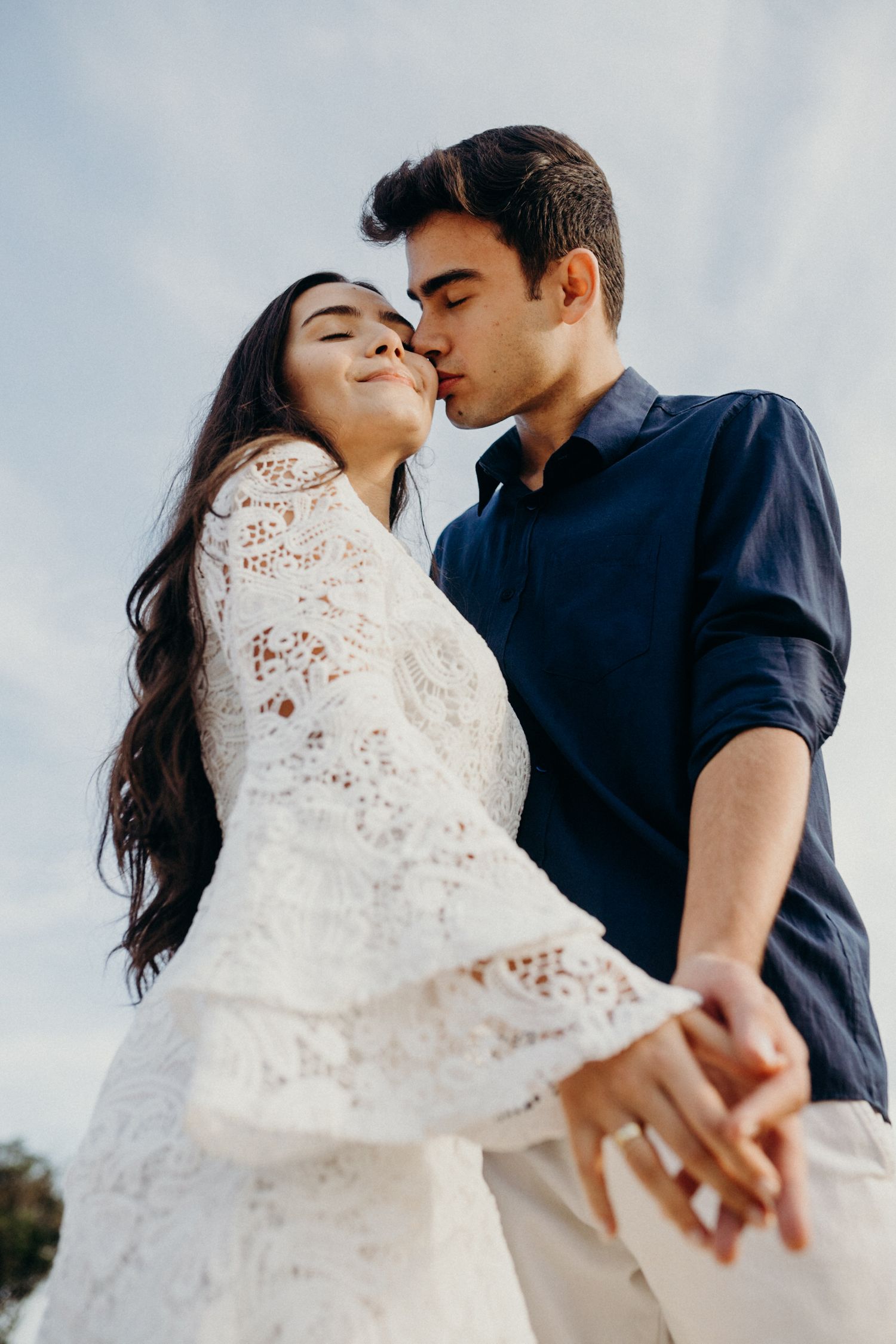 A tender moment captured between two people wearing white and navy blue, holding hands against a bright sky.