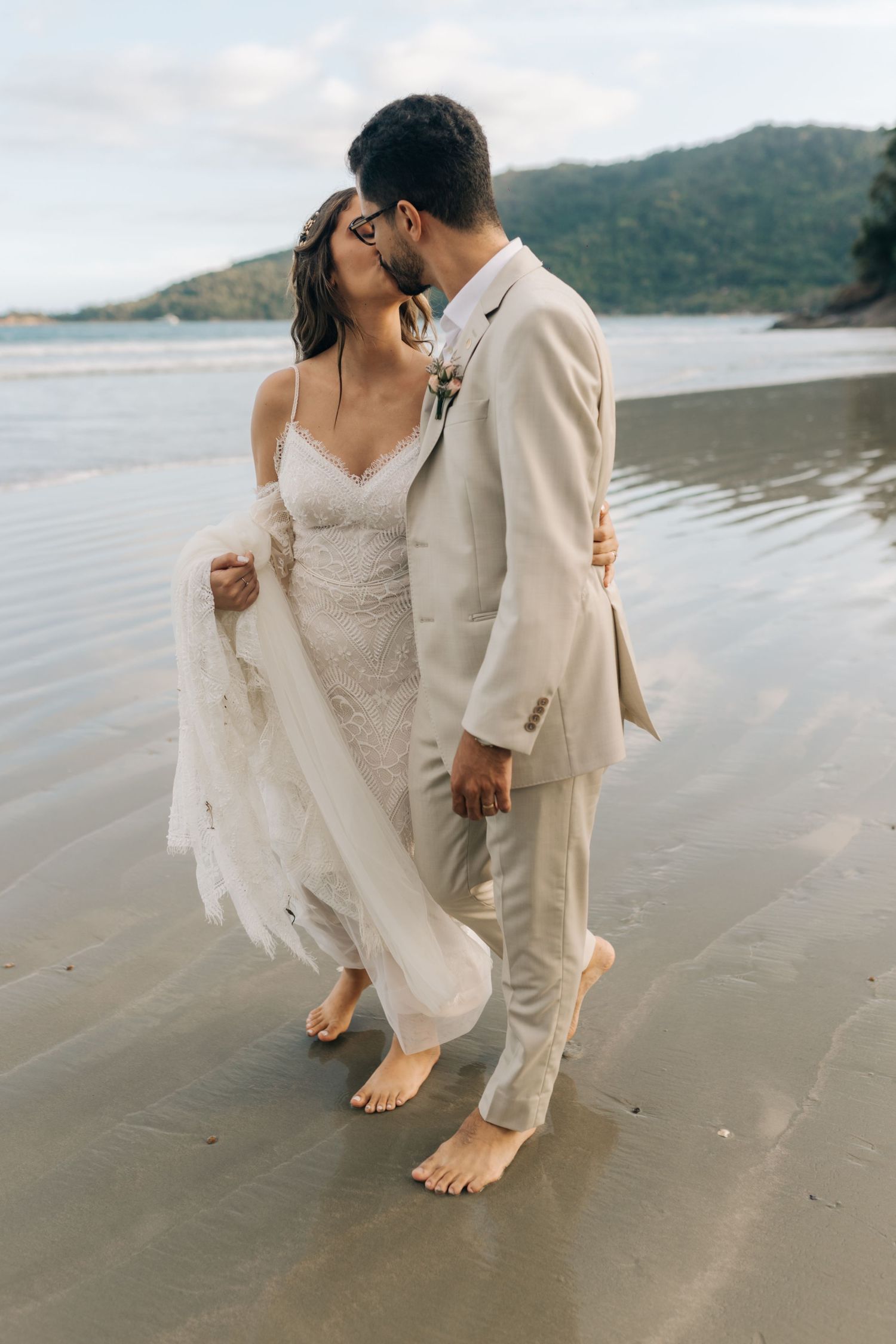 Intimate beachside wedding moment with barefoot newlyweds in elegant white dress and beige suit.