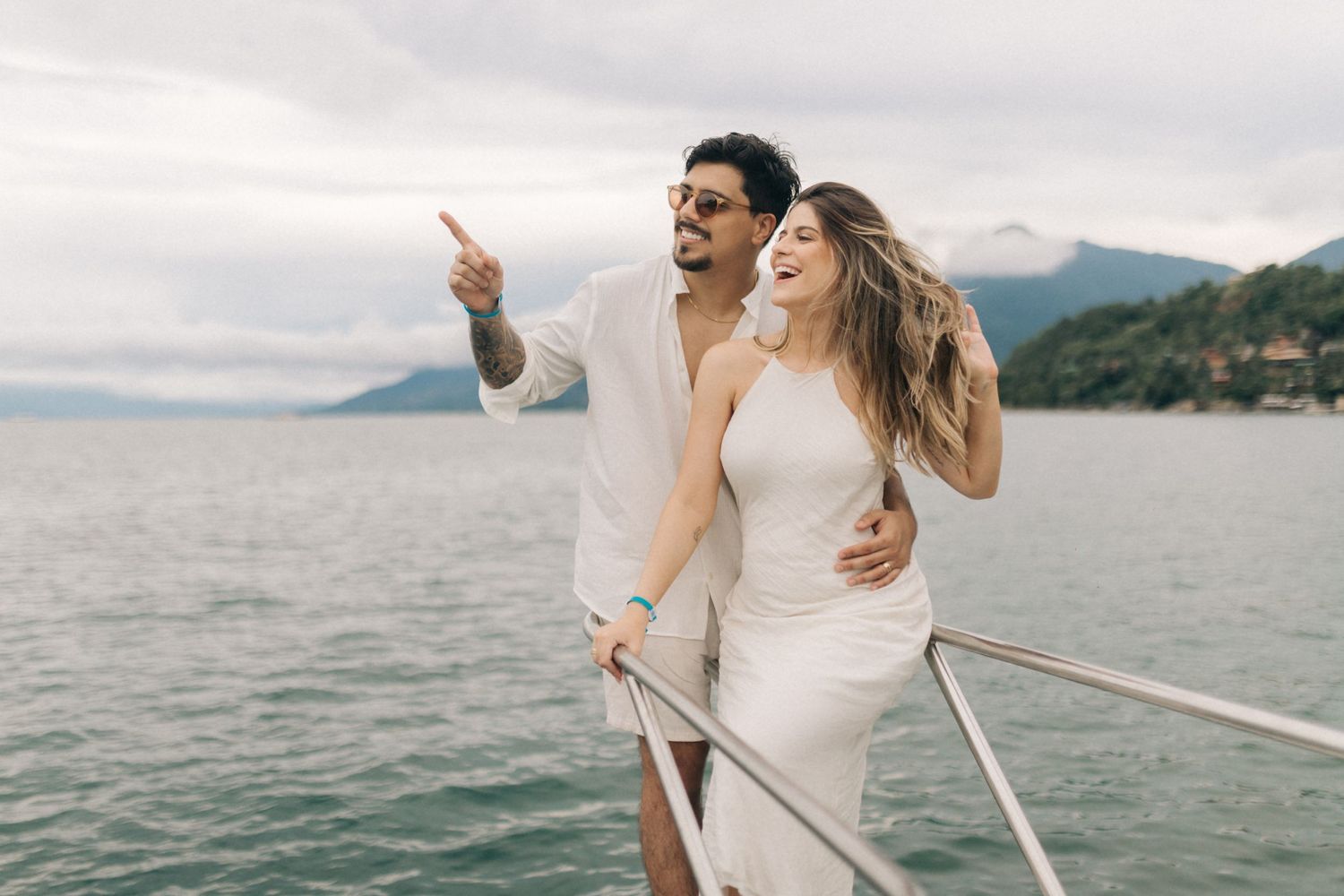 Happy couple pointing and enjoying a moment together on a boat with mountainous landscape behind them.