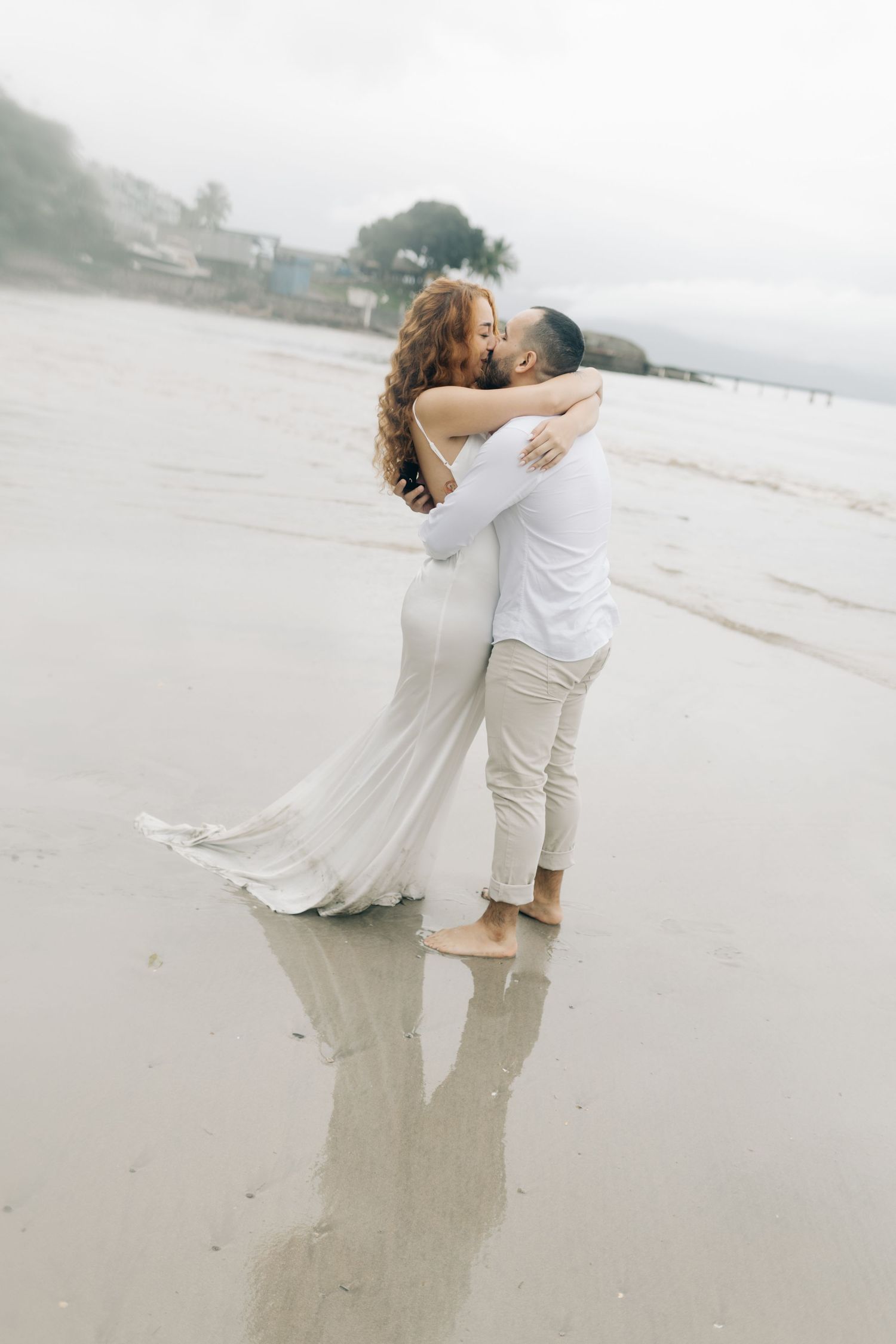 A couple in matching white outfits dances together on a reflective wet beach during an overcast day.