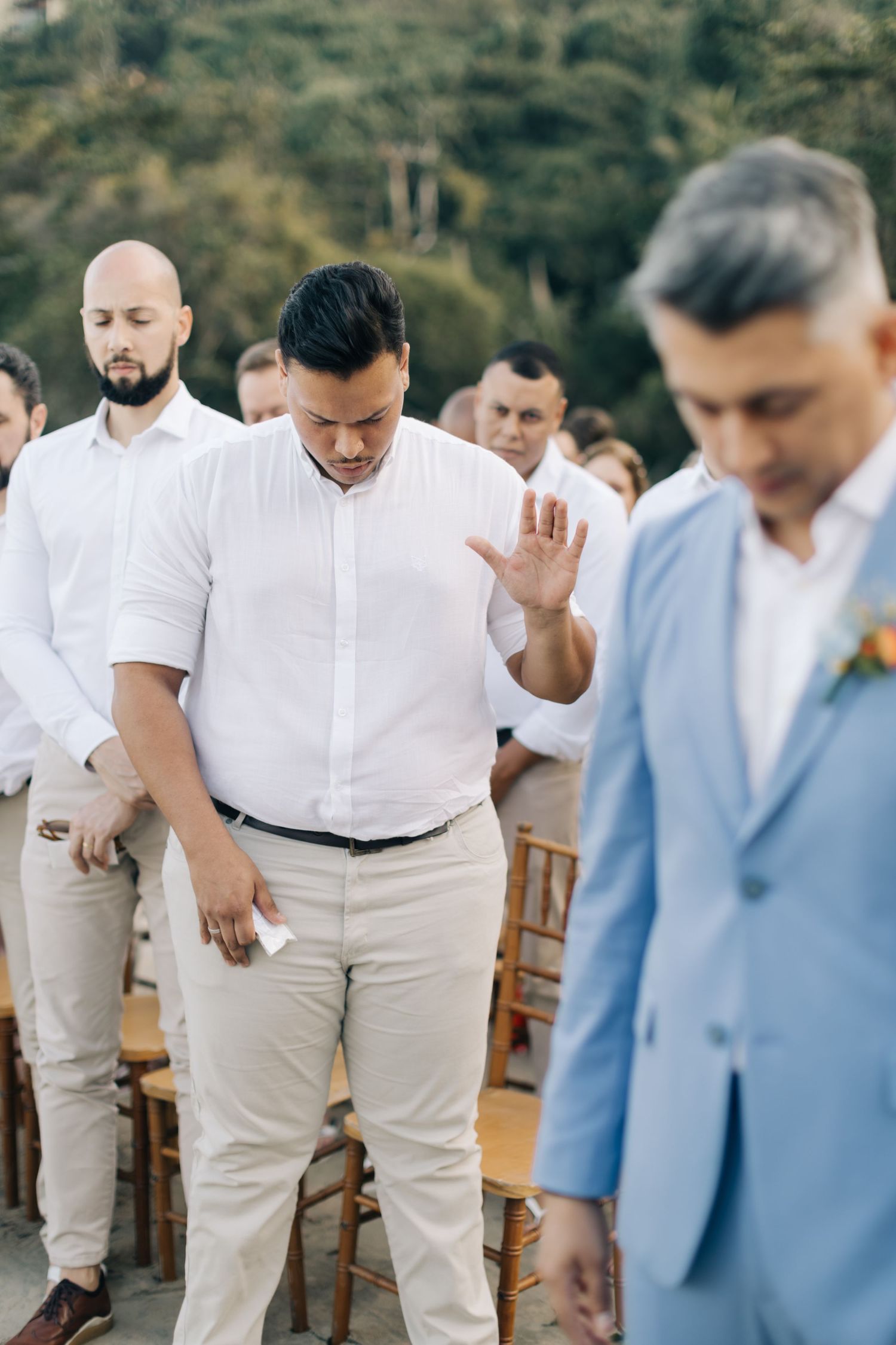 Group of wedding guests in light colored attire participate in prayer during outdoor ceremony.