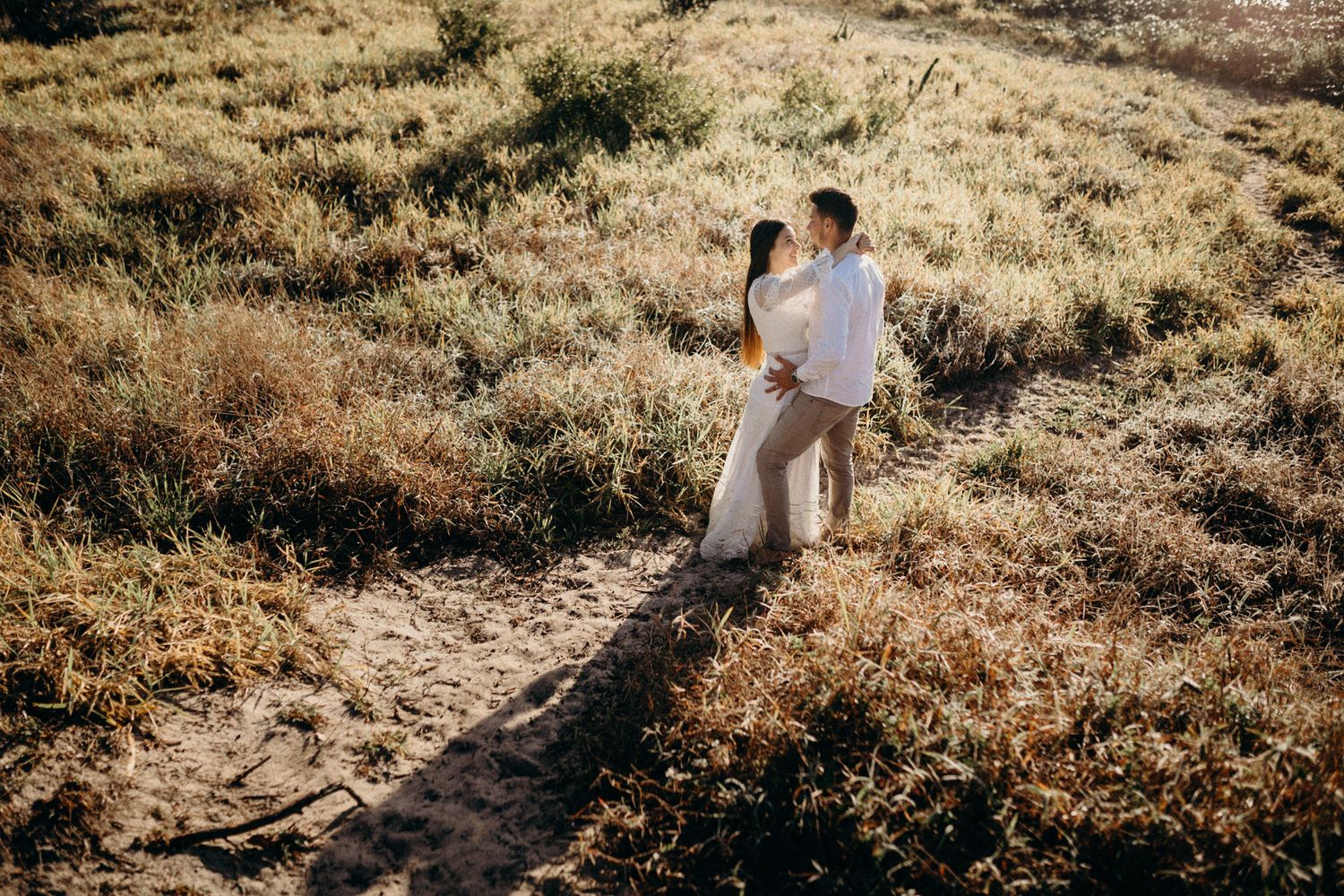 Romantic couple embraces on a hilltop path surrounded by golden desert brush and warm sunlight.