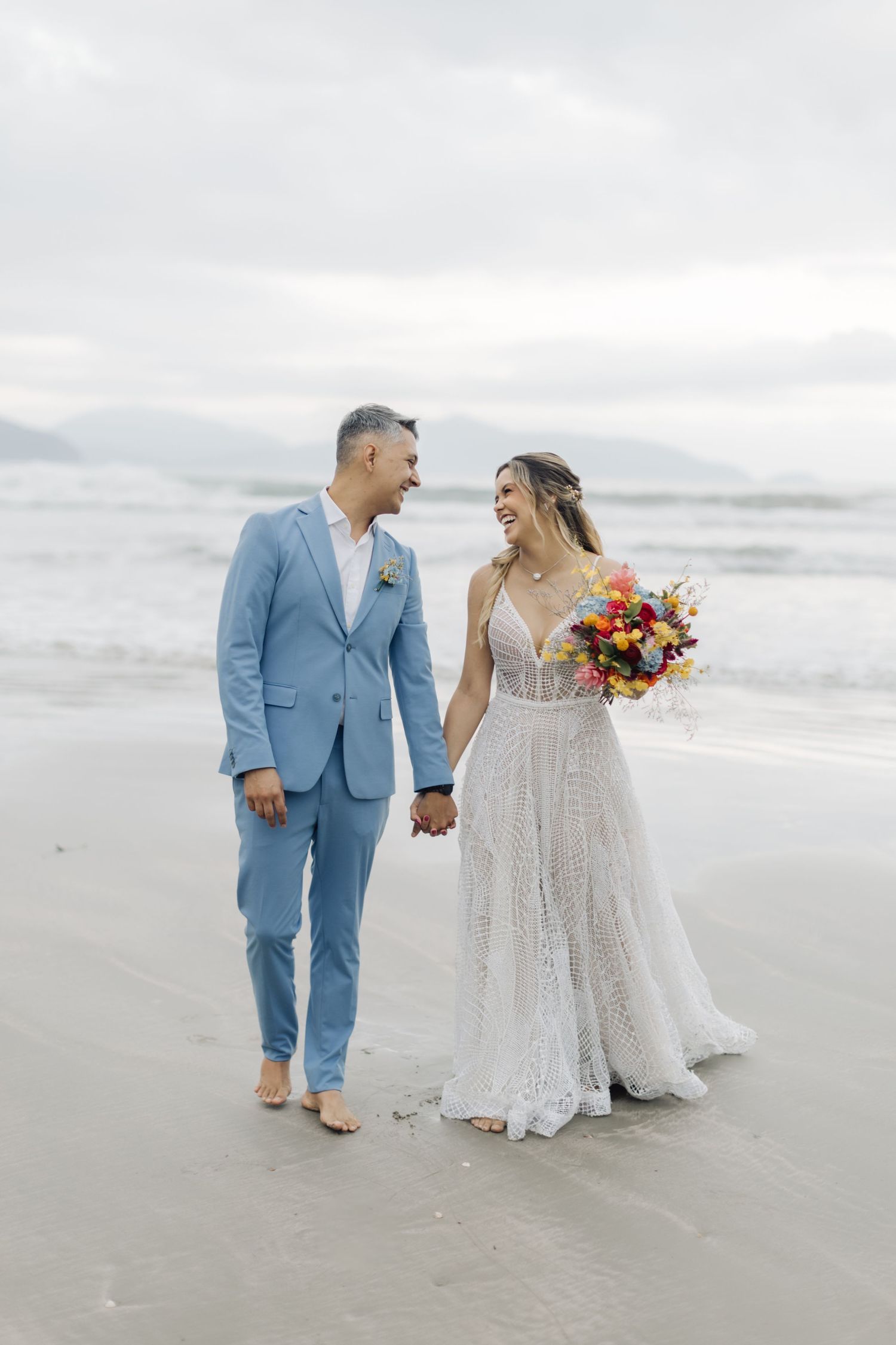 Couple walking hand in hand on a cloudy beach with the bride holding a colorful bouquet.