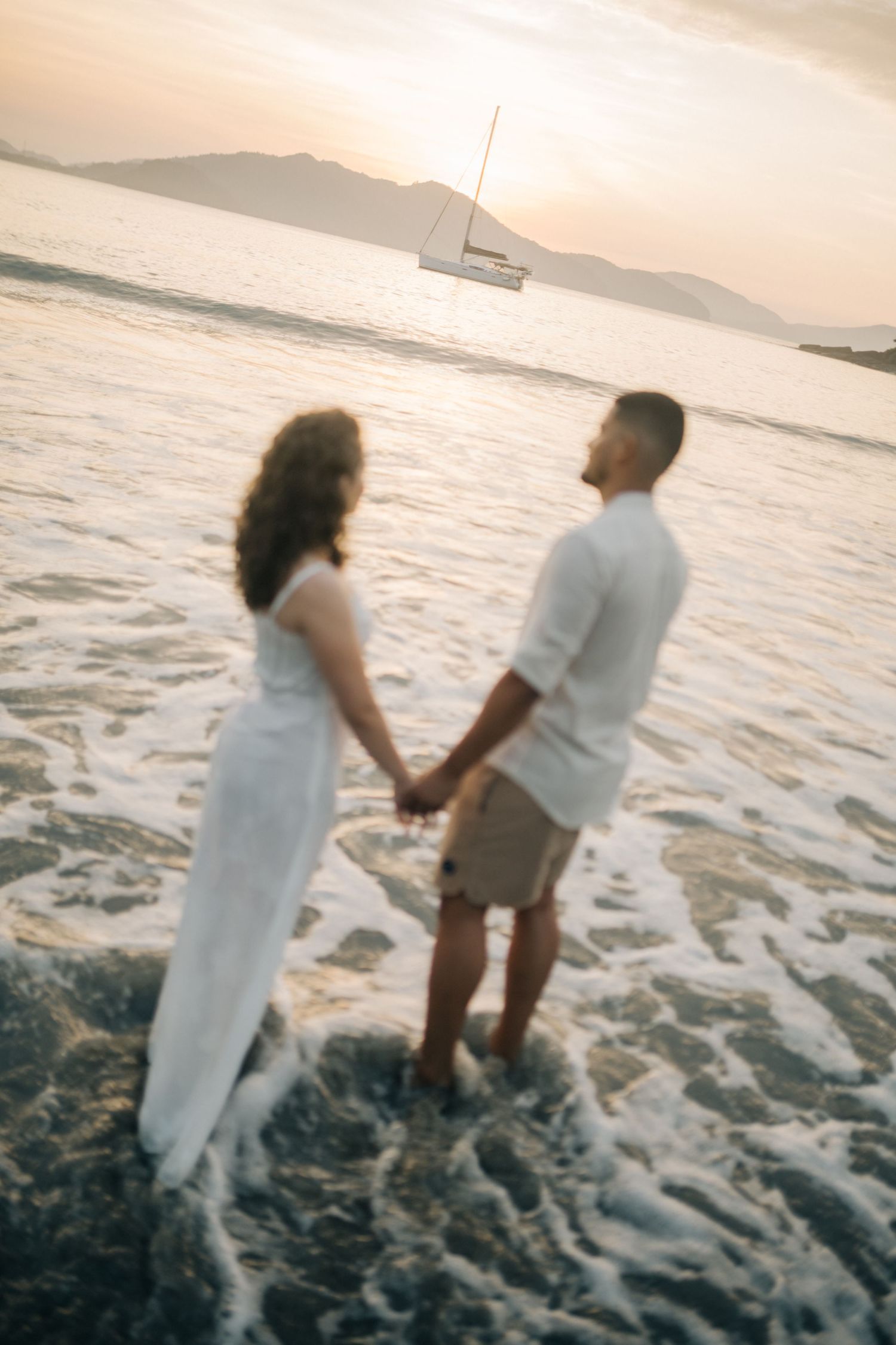 Romantic couple holding hands at sunset on a beach with a sailboat in the distance.