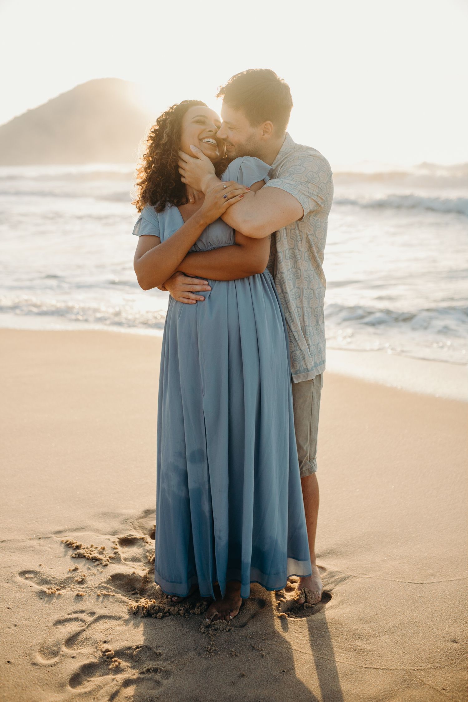 Romantic couple embracing on beach at sunset with waves and mountains in background.