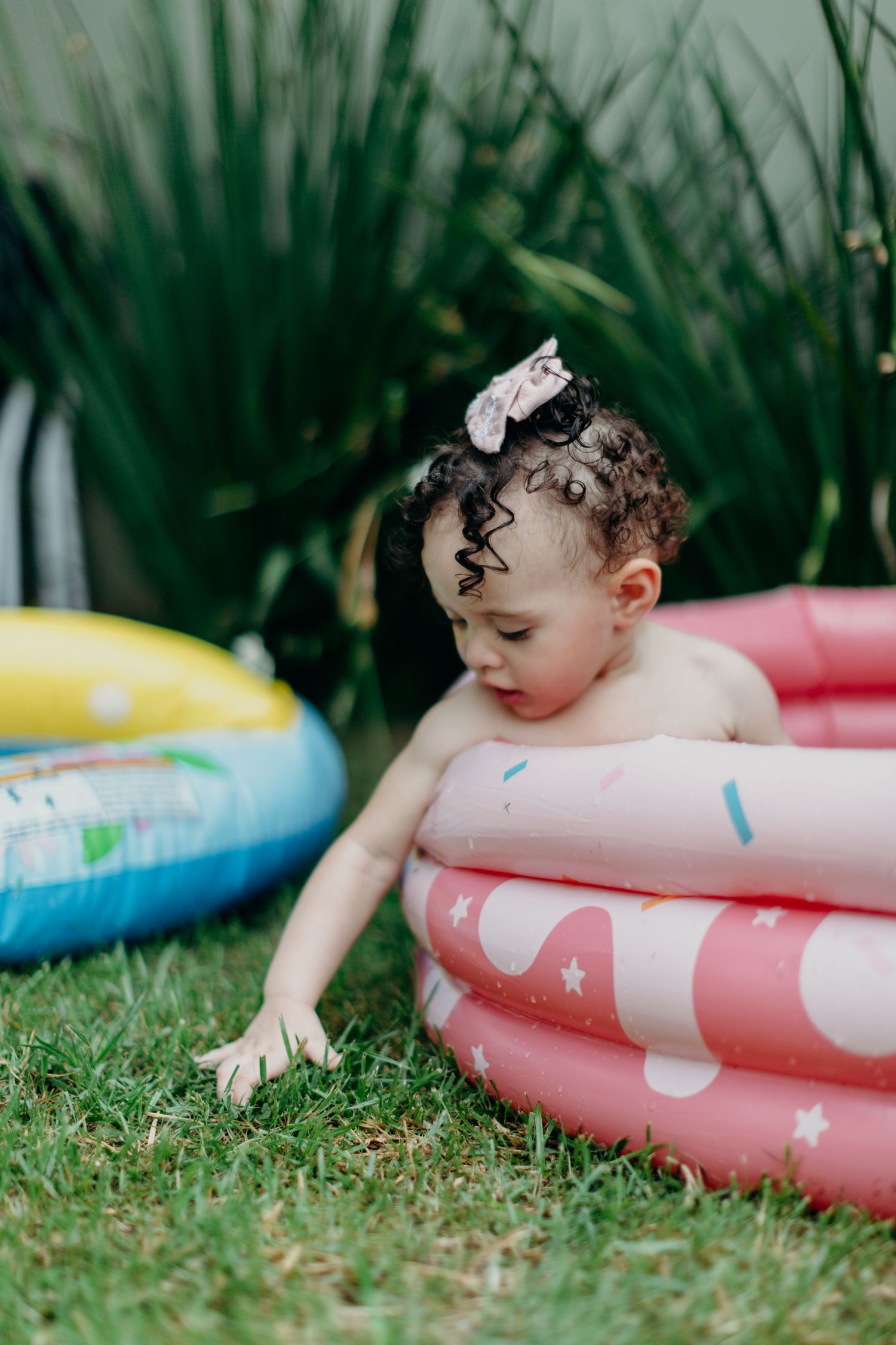 A toddler plays in a colorful inflatable pool on a grassy lawn during summer.