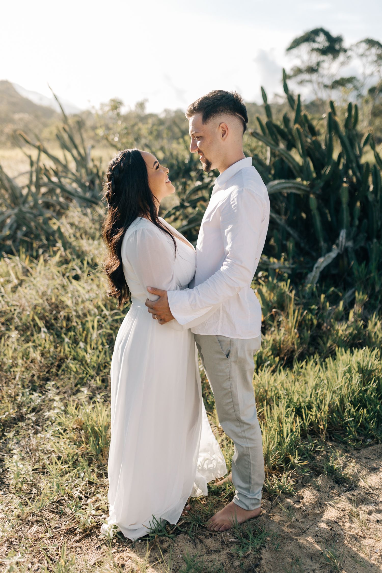 A couple in matching white outfits share an embrace on a hillside path during golden hour.