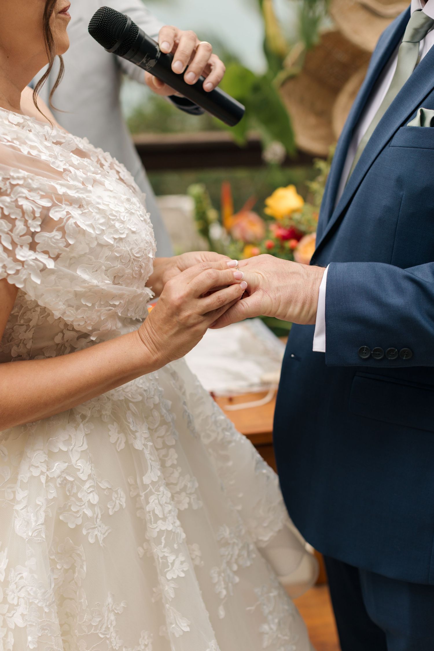 Close-up of hands exchanging rings during wedding ceremony with lace dress details visible.