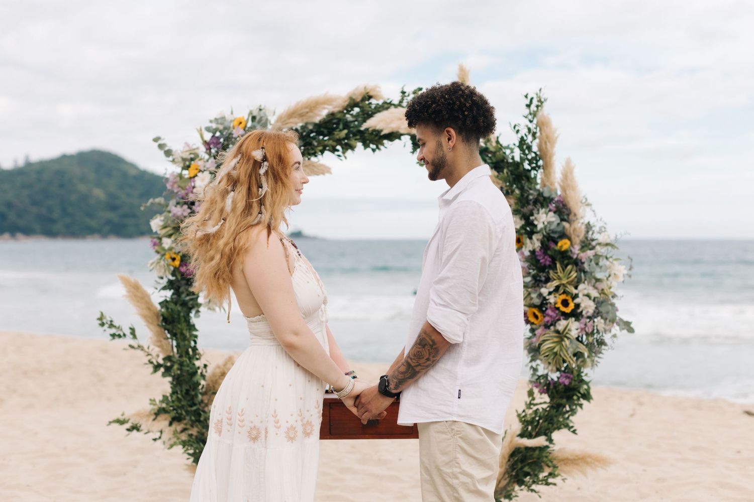 Couple holds hands under a flower-adorned arch during their seaside ceremony.