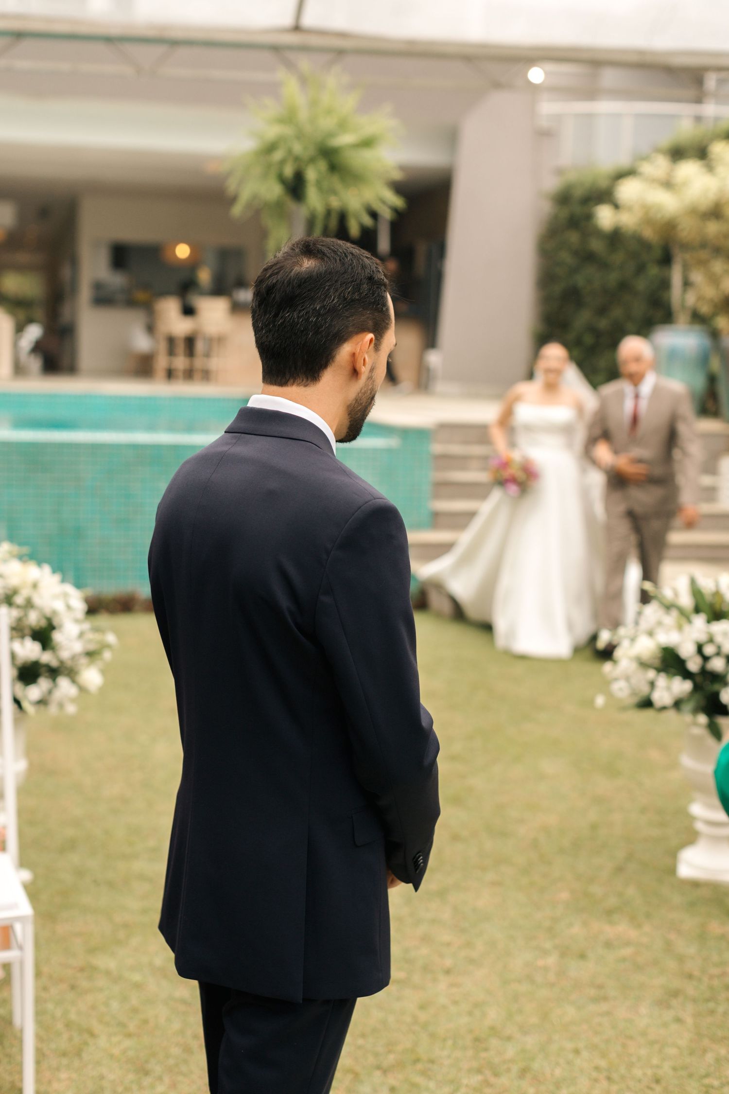 A groom in a dark suit watches the processional from across a pool deck decorated with flowers.