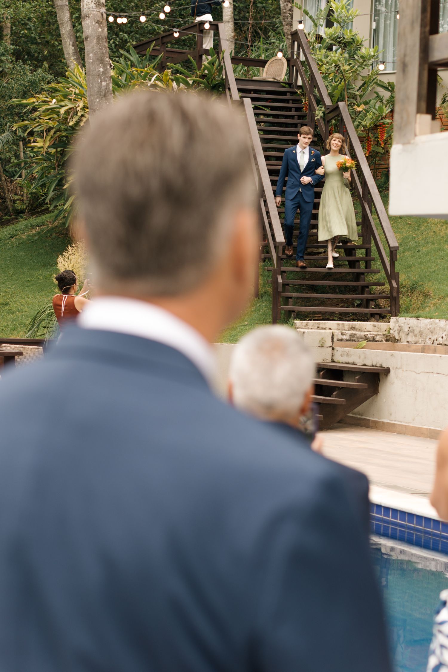 View from behind guests watching a wedding ceremony by a pool with string lights overhead.