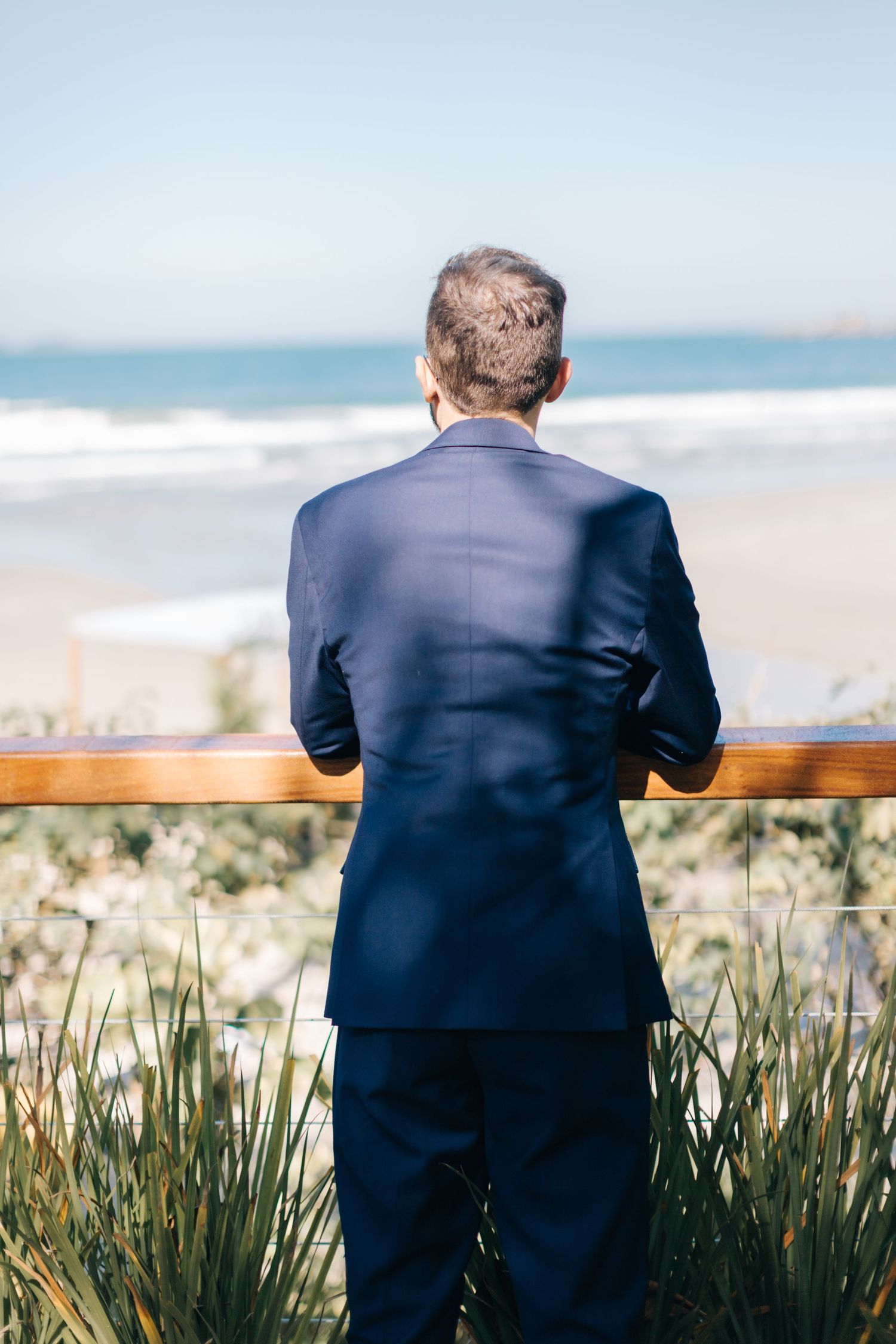 Figure in dark suit looks out over ocean view from wooden deck railing.