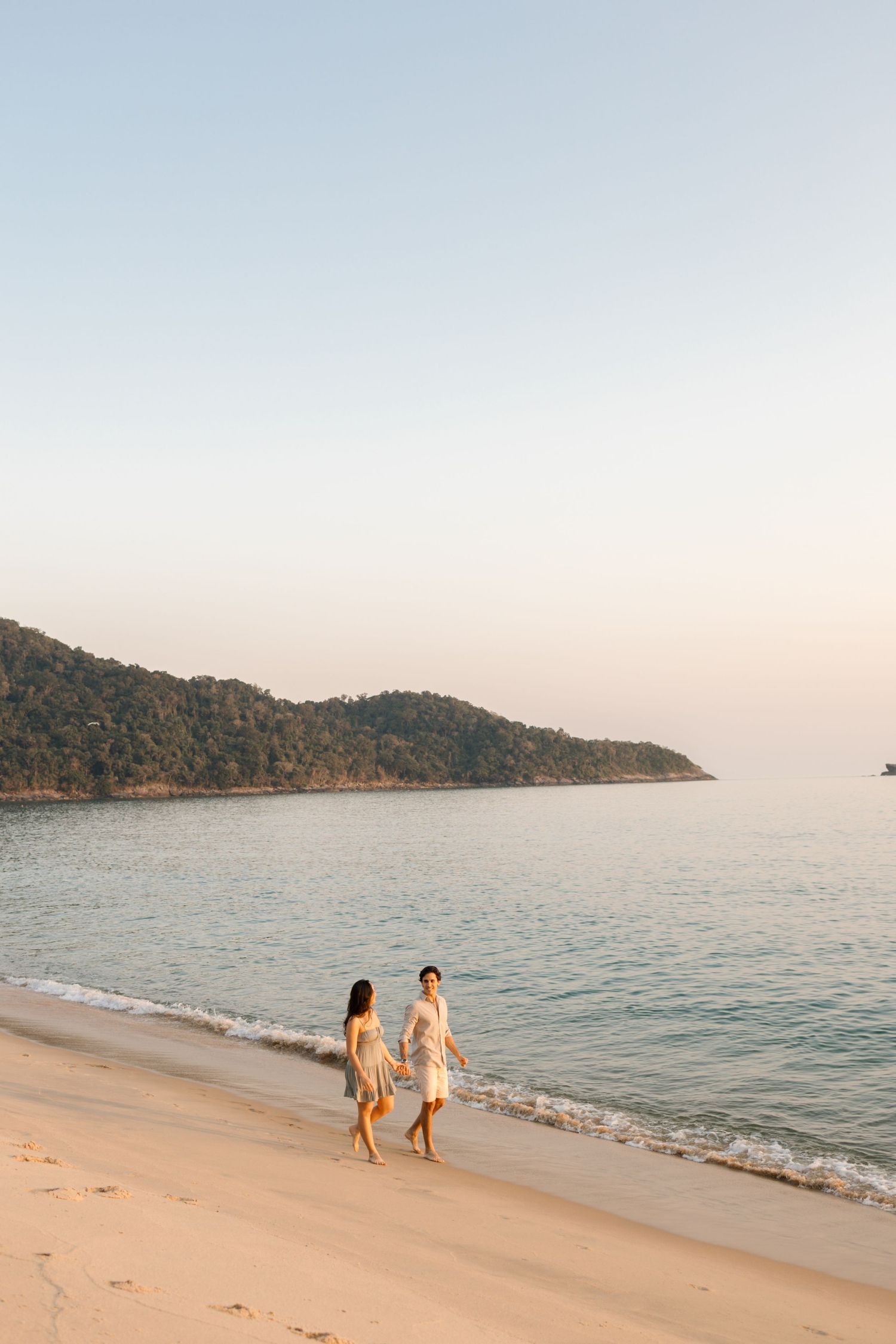 People walking along a serene beach shoreline at sunset with mountains in the background.