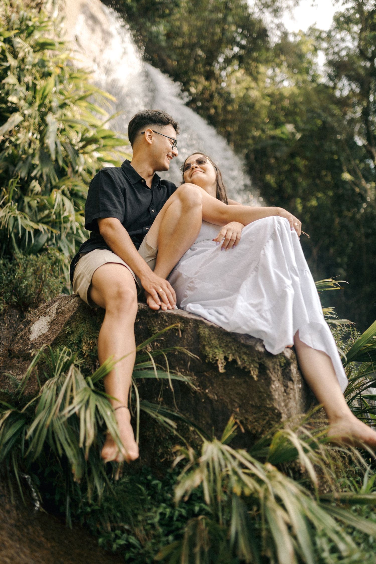 Intimate couple's photoshoot on a rocky ledge near a flowing waterfall in a tropical setting.