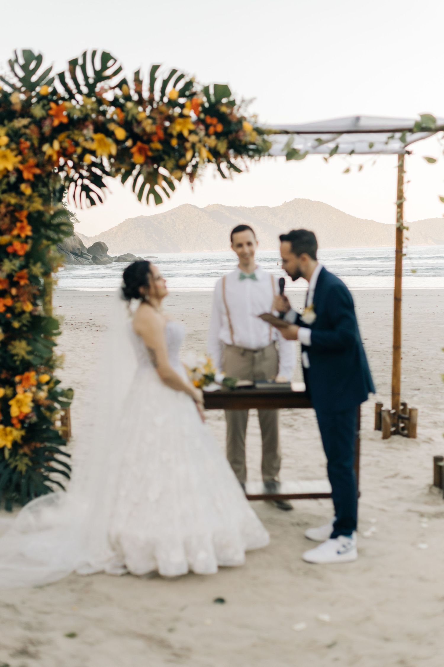 Beach wedding ceremony under floral arch with mountains in background.