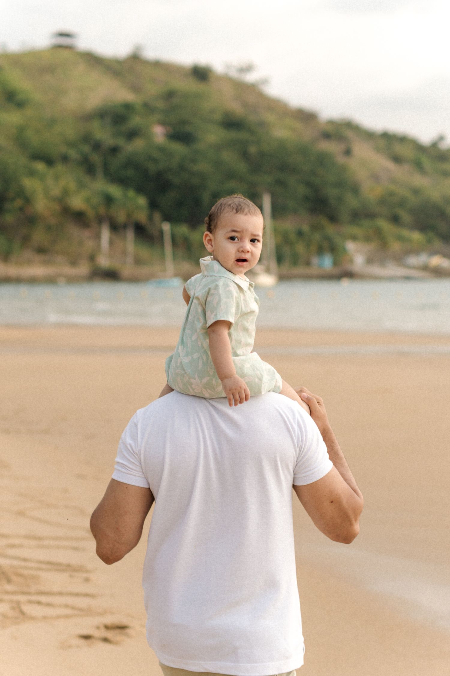 A baby rides on someone's shoulders while walking along a scenic beach with hills in the background.