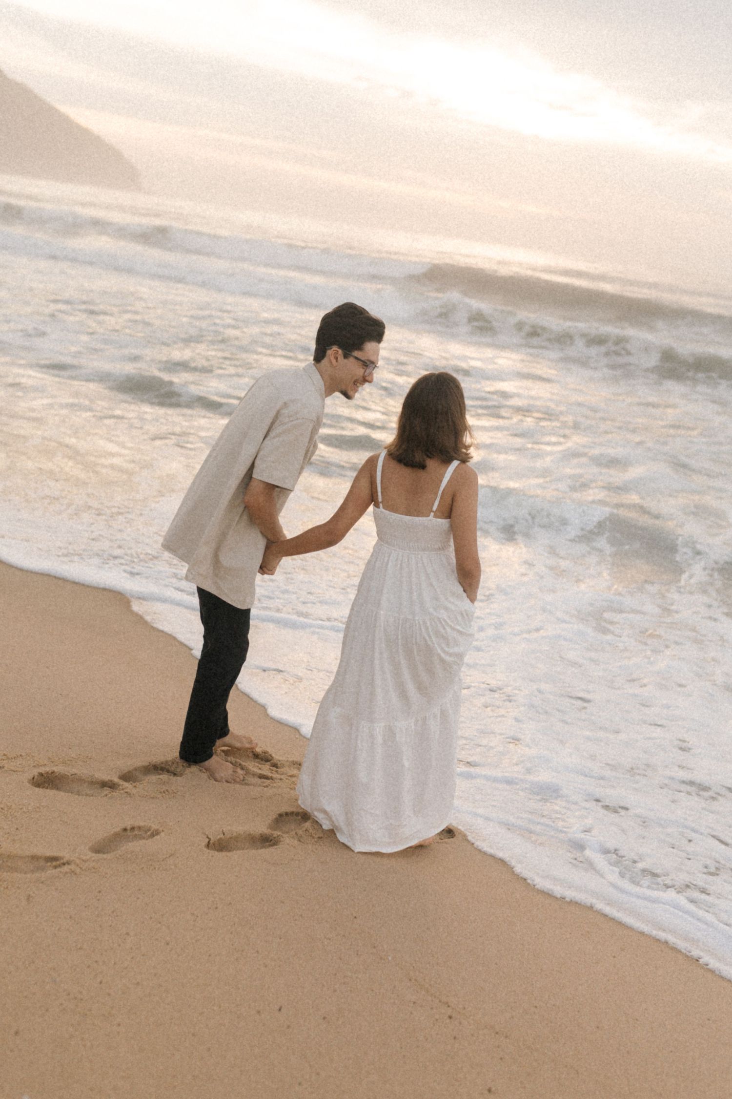 A romantic couple strolls along a scenic beach during golden hour with waves crashing behind them.