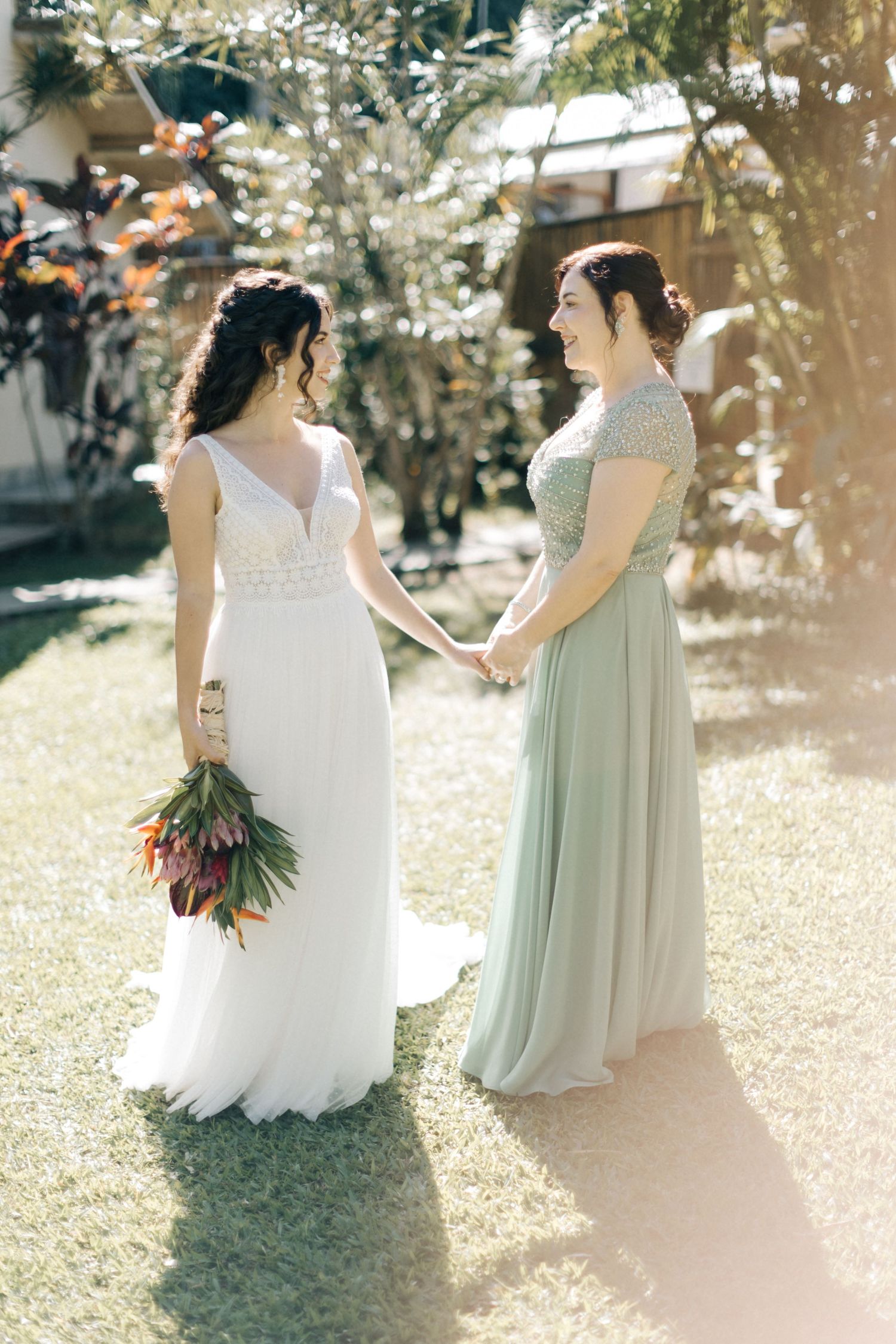 Two people in wedding attire standing together in a sunlit garden with tropical flowers.
