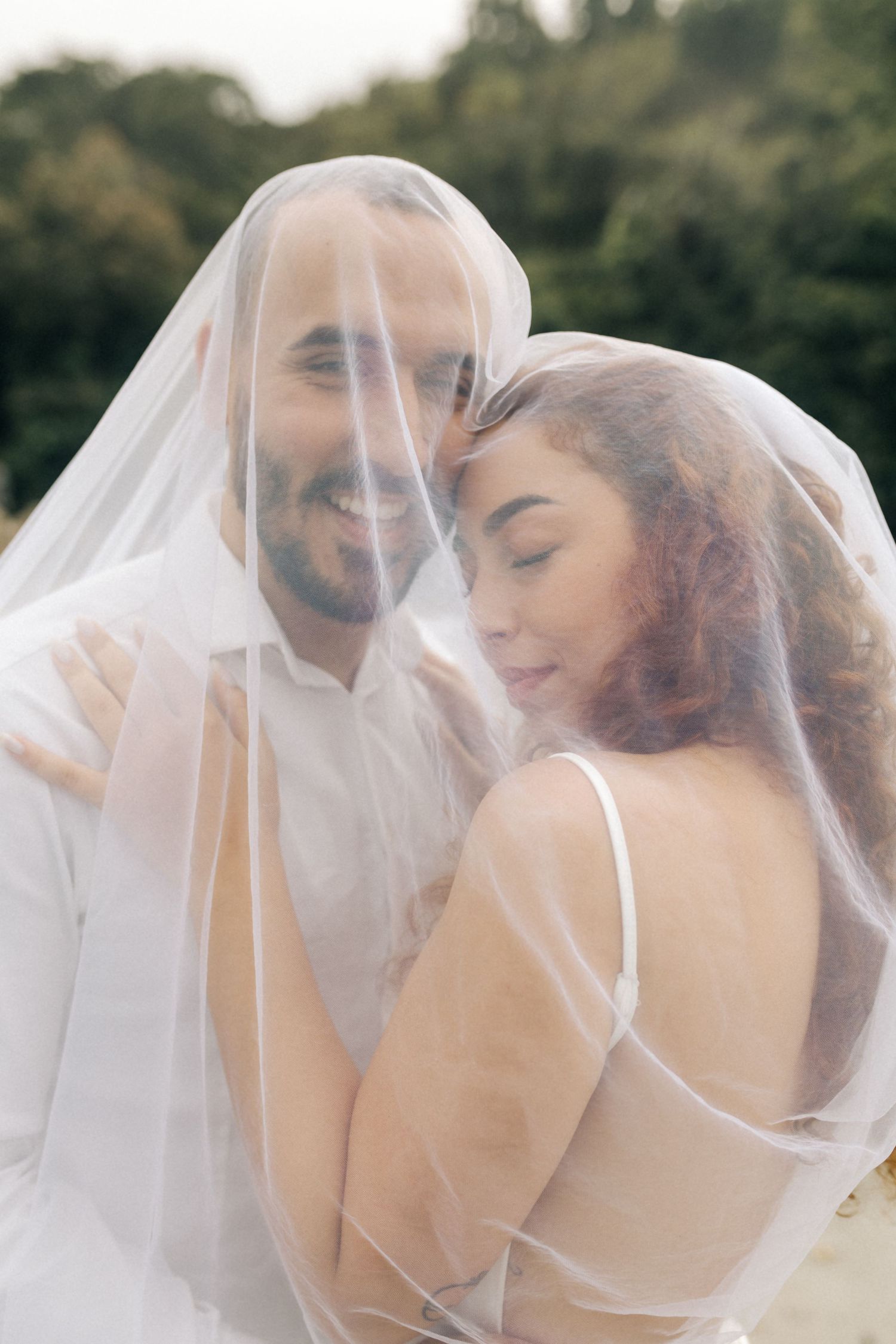 Romantic couple embracing under sheer wedding veil with soft natural lighting.