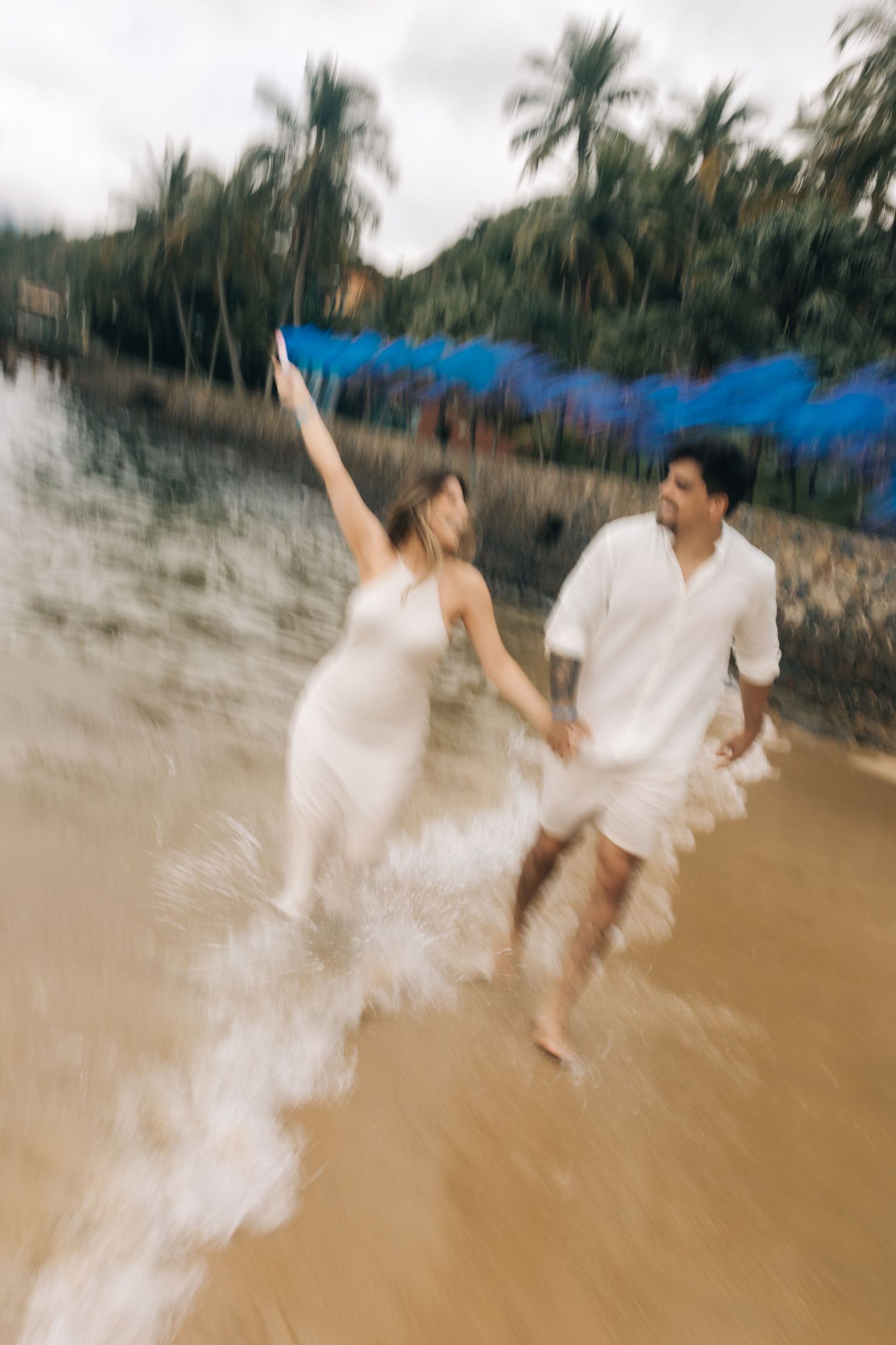 A couple in white outfits runs playfully through shallow water on a tropical beach with blue smoke effects.